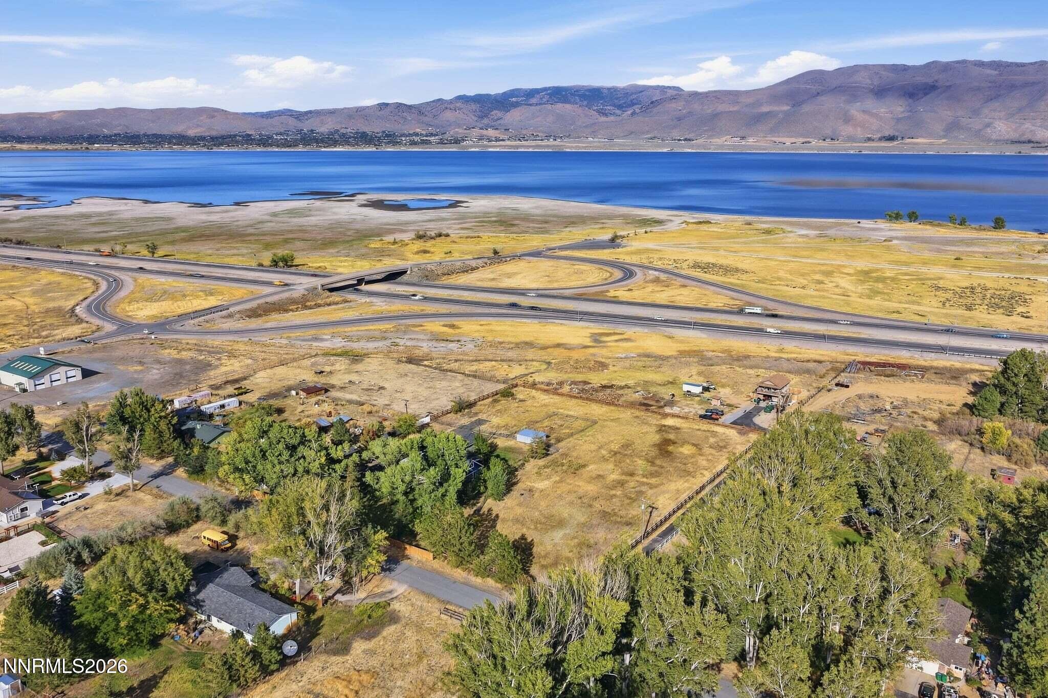 Aerial view of homes and dry fields beside a lake, with a highway interchange and mountains beyond.
