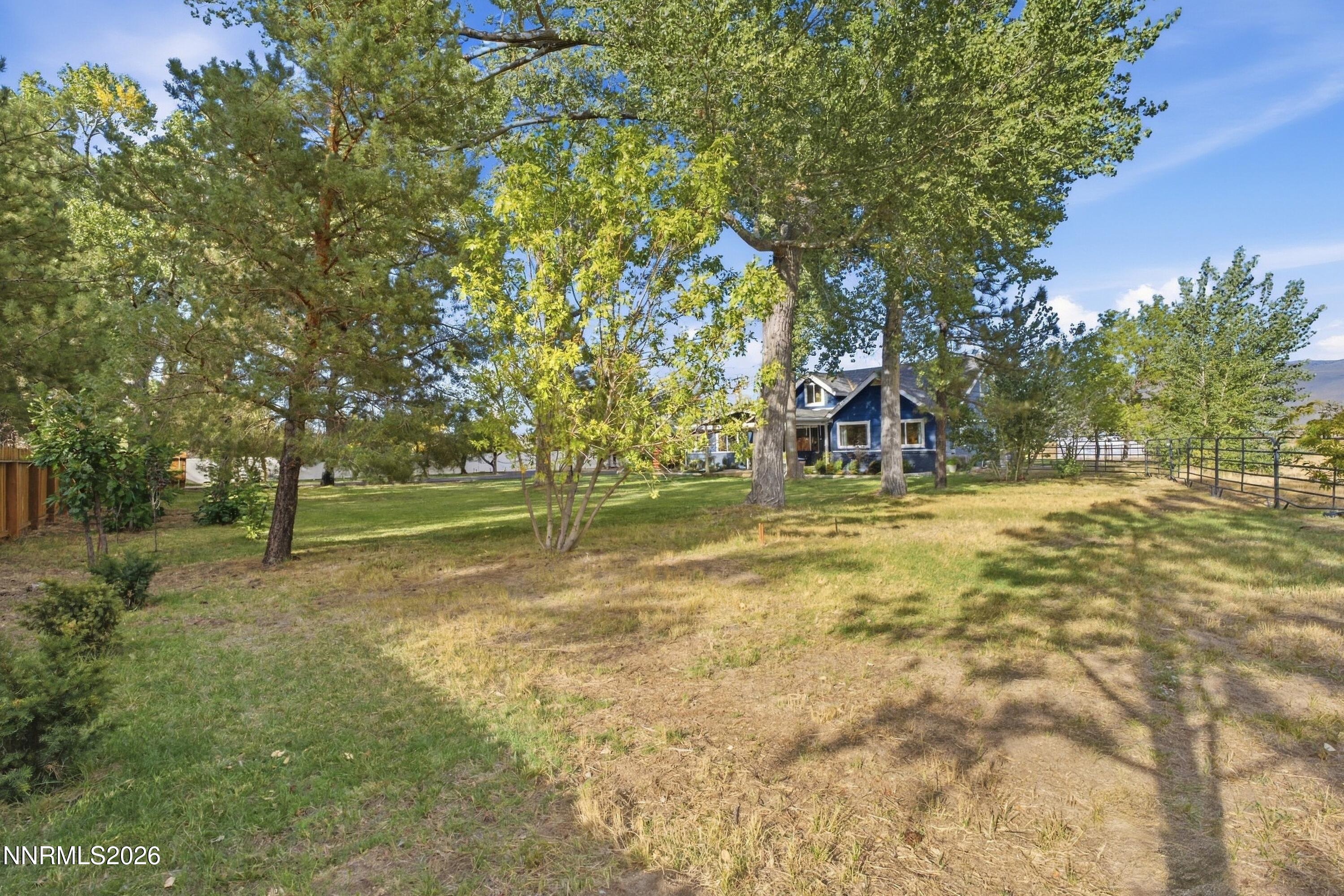 Blue house set among mature trees on a spacious yard, with a metal fence and distant hills.