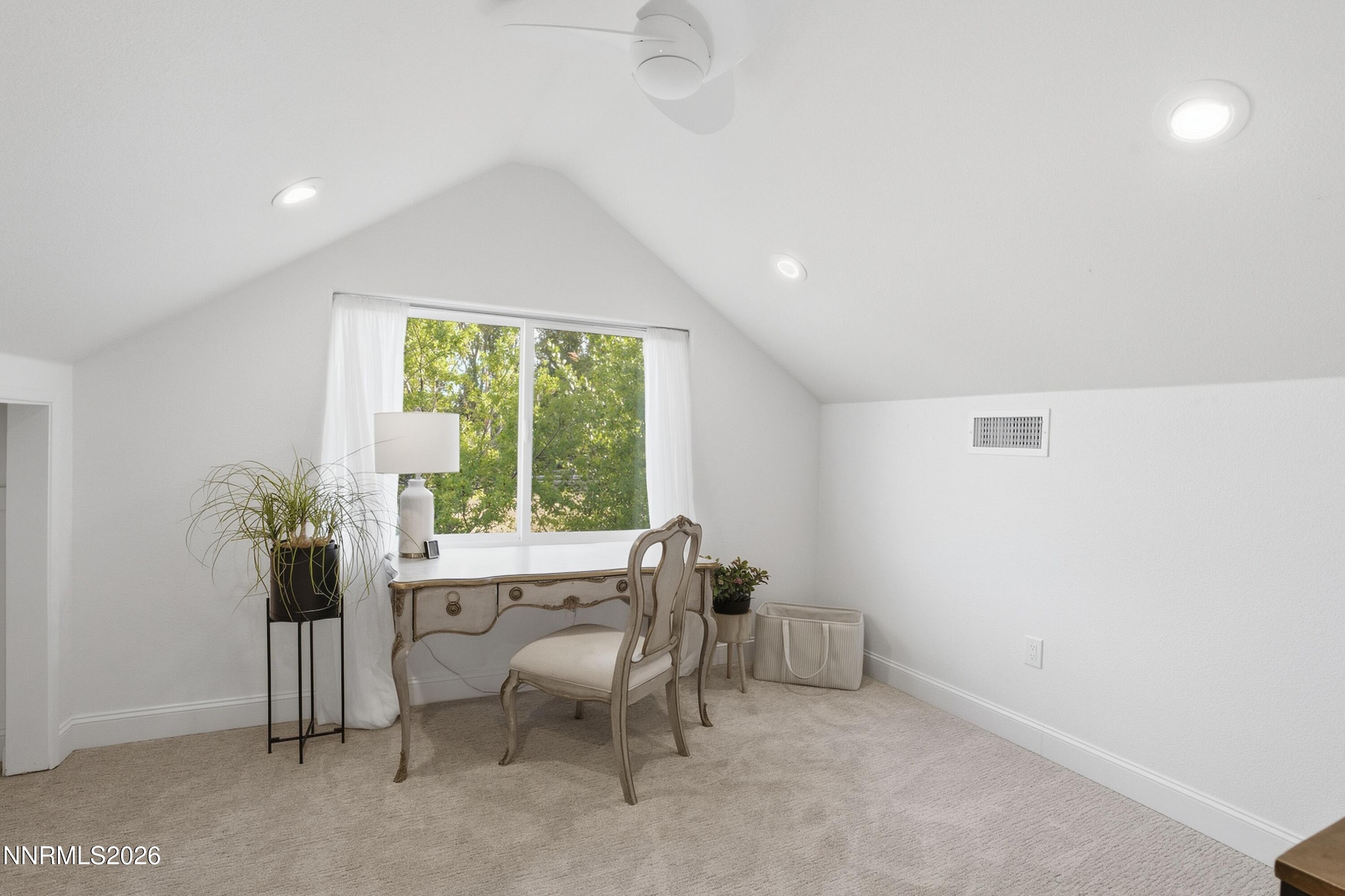 Staged home office with a desk and chair beneath a sloped ceiling, beside a window overlooking leafy trees.