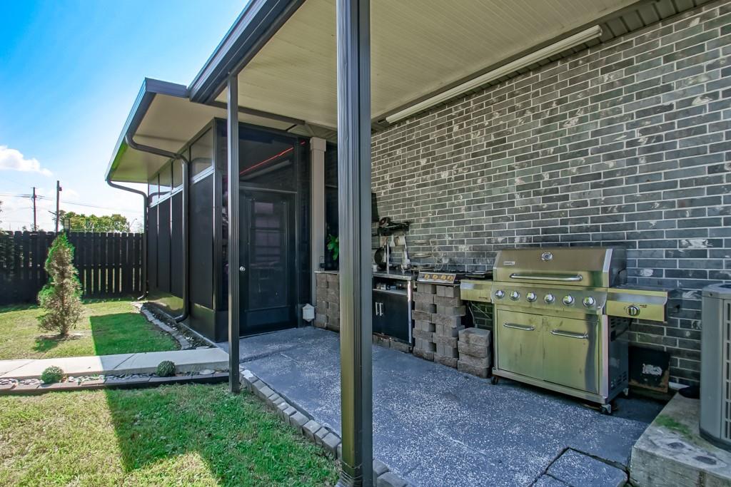 Covered patio with a stainless steel gas grill beside a small fenced lawn.
