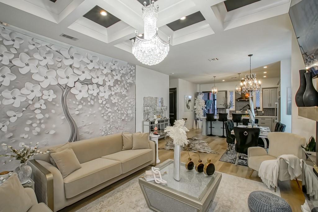 Living room with a beige sectional, sculptural floral wall, and crystal chandeliers opening to the kitchen.