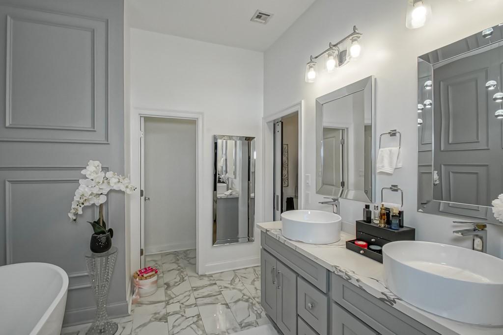 Bathroom with two vessel sinks, gray paneled cabinetry, marble-patterned surfaces, and a freestanding tub.
