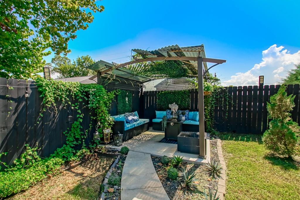 Backyard patio with blue-cushioned seating beneath a pergola, surrounded by greenery and a black privacy fence.