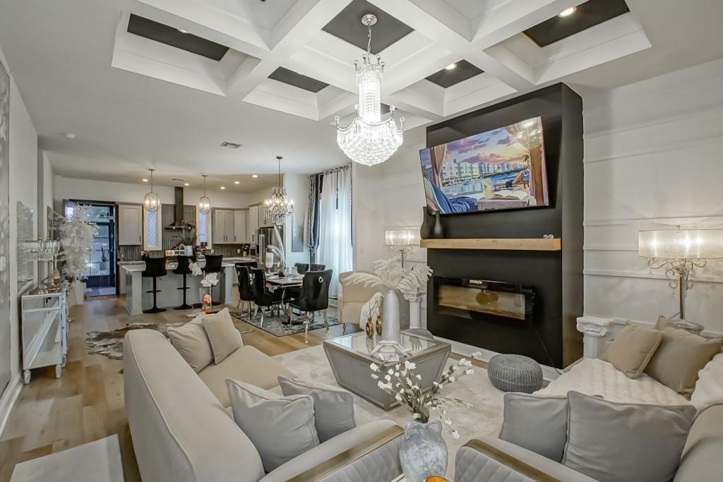 Open-plan living room with a coffered ceiling, crystal chandelier, and black fireplace.