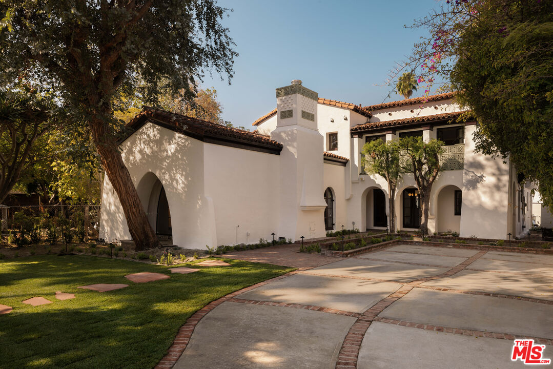 White stucco Spanish-style home with arched openings, a tiled roof, and a tree-shaded courtyard.