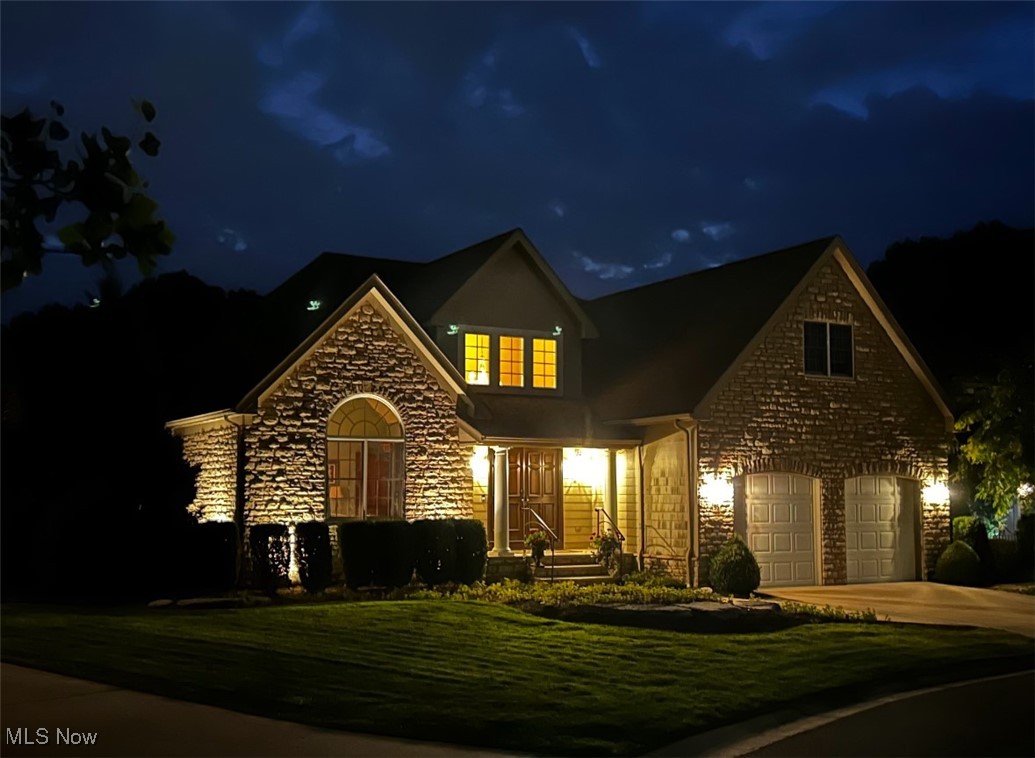 Two-story stone-and-shingle home at night, with an arched window, illuminated entry, and two-car garage.