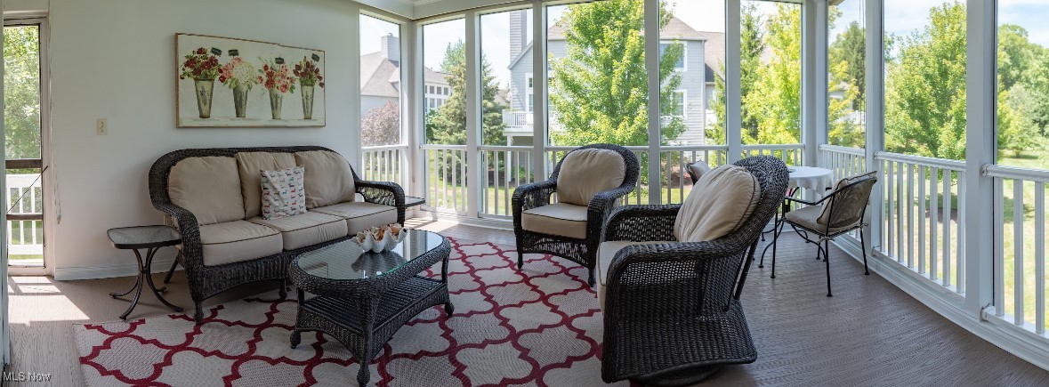 Furnished sunroom with wicker seating, patterned rug, and floor-to-ceiling windows overlooking trees.