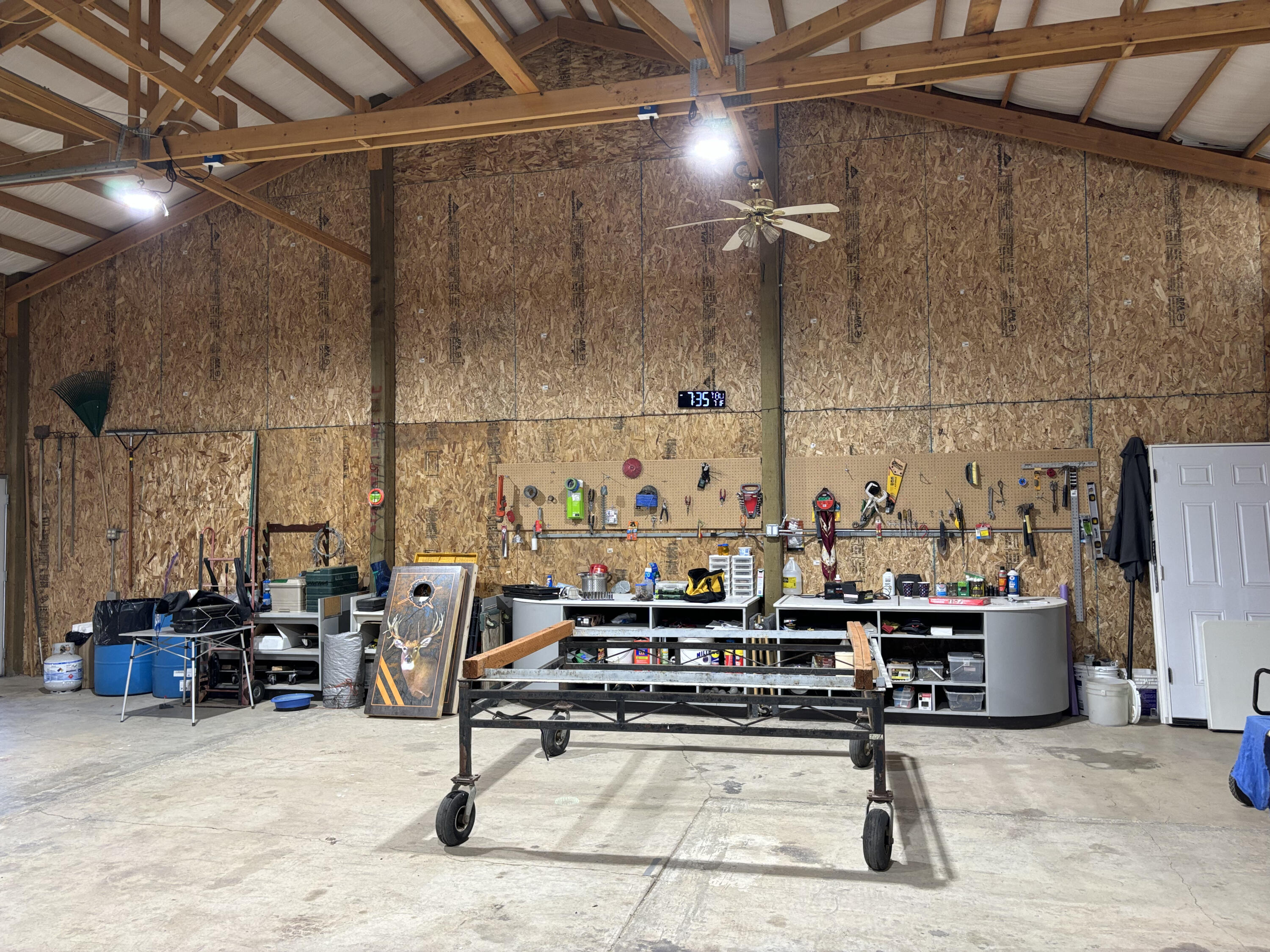 Workshop with exposed roof trusses, tool-covered pegboards, and a wheeled work stand.