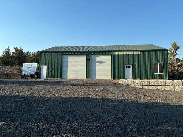Green metal outbuilding with two large garage doors, a gravel forecourt, and a low stone retaining wall.