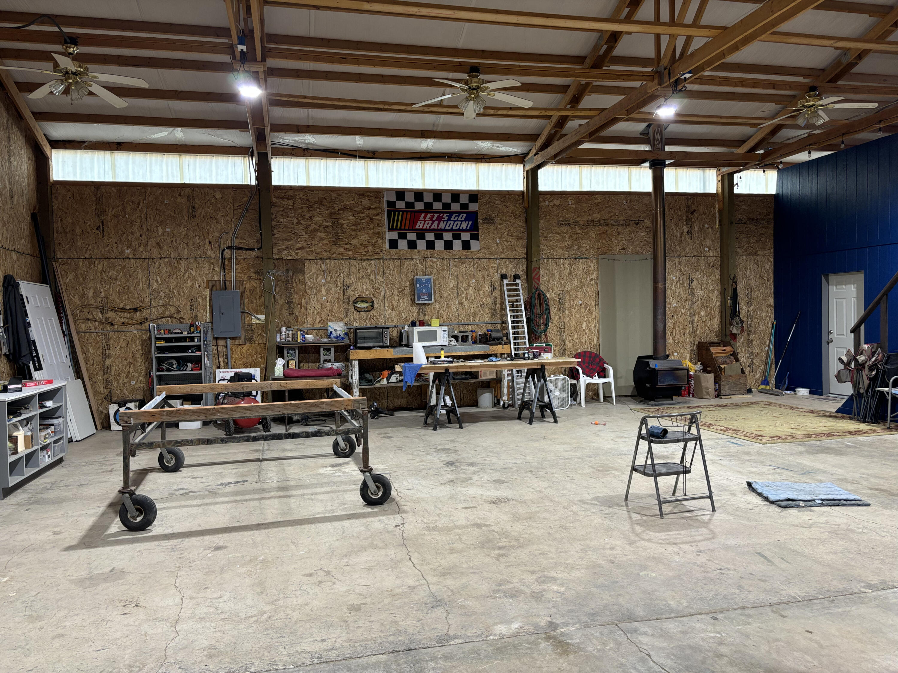 Large workshop with exposed wood framing, workbenches, concrete floor, and a blue wall with a white door.