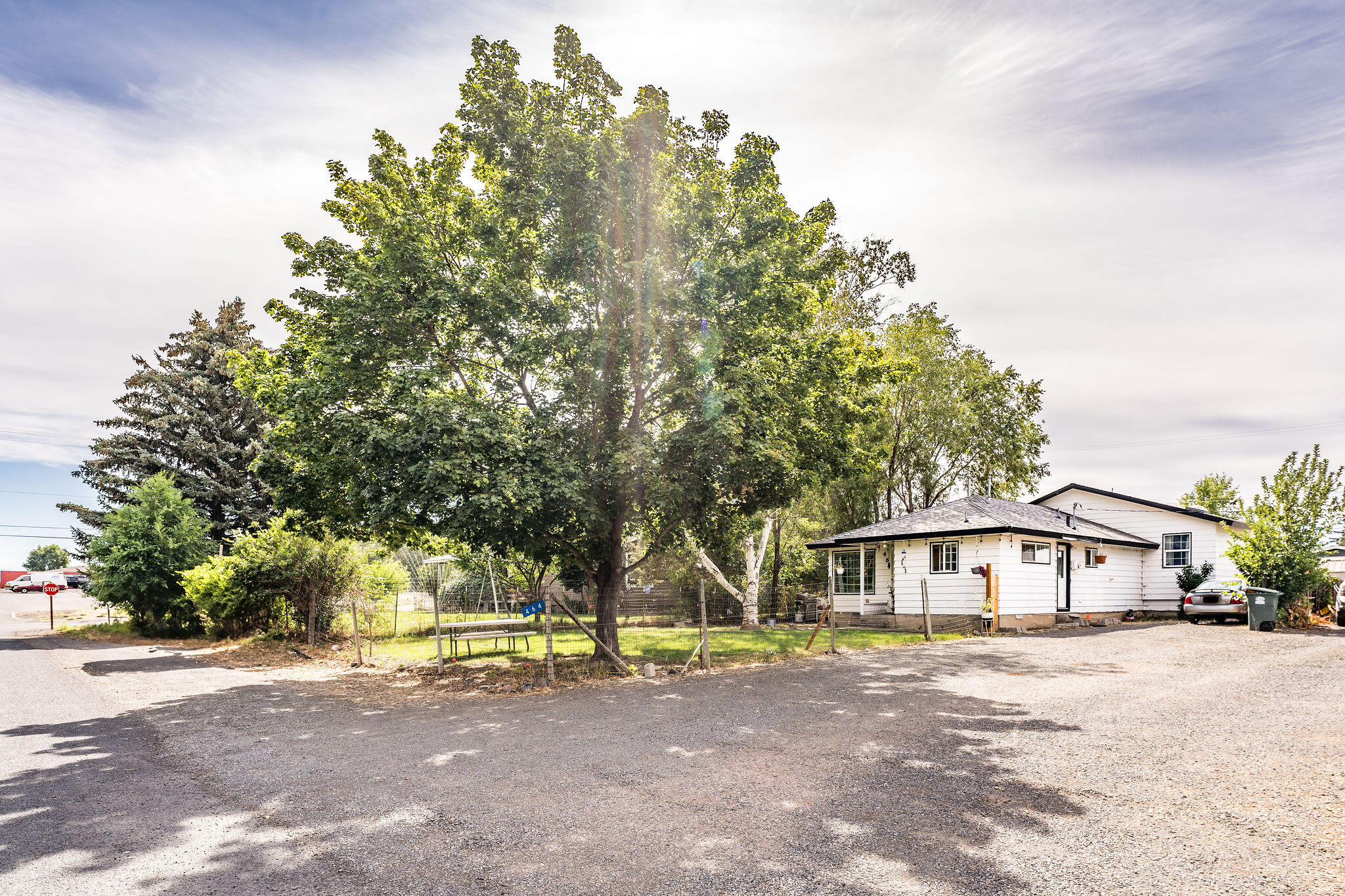 White single-story house beside a gravel driveway, with a fenced lawn and large shade tree.