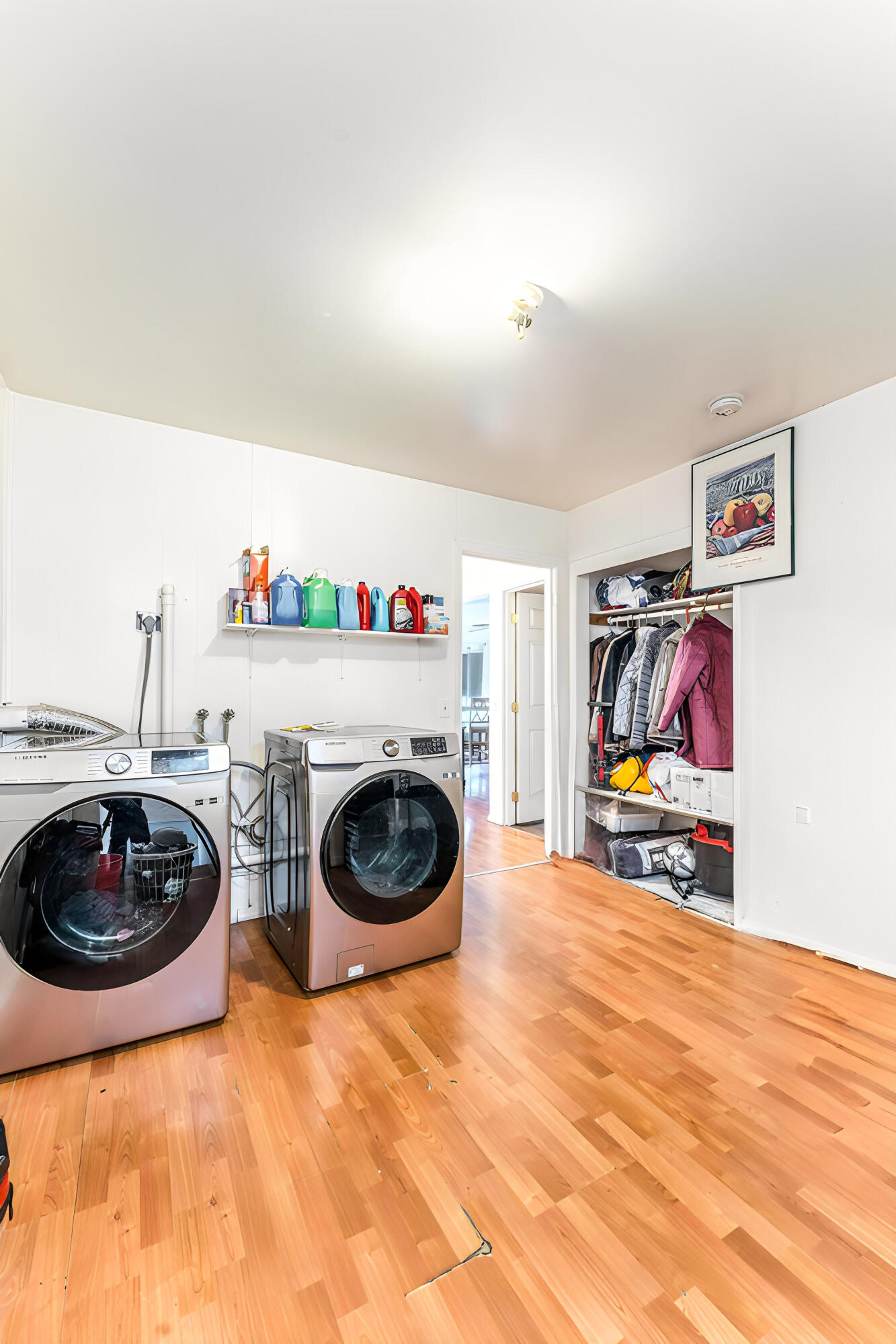 Laundry room with two front-loading machines, a supply shelf, and an open closet of coats and bags.