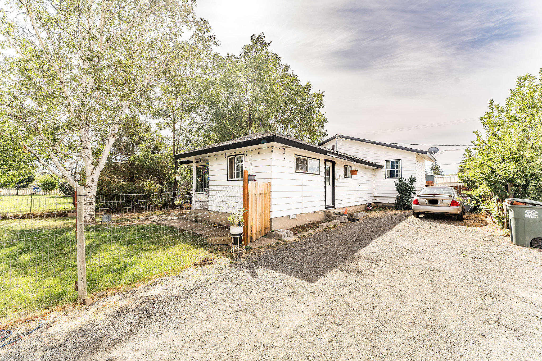 White single-story house beside a gravel driveway, parked car, and fenced, tree-shaded yard.
