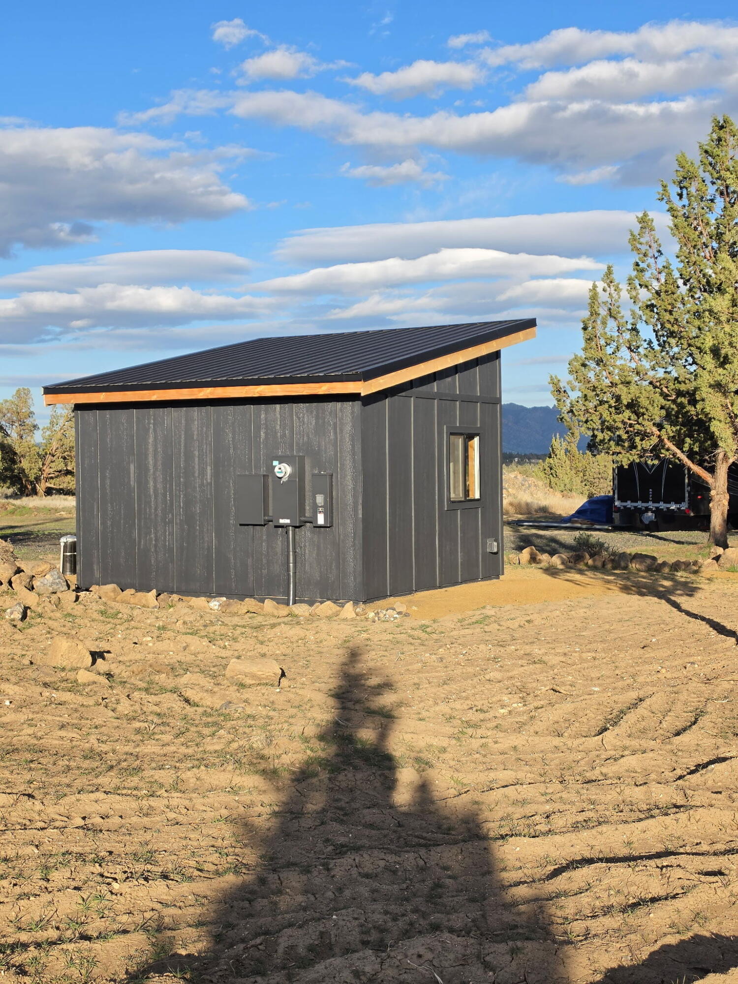 Small charcoal outbuilding with a sloped metal roof on a dry, rocky lot, with trees and mountains beyond.