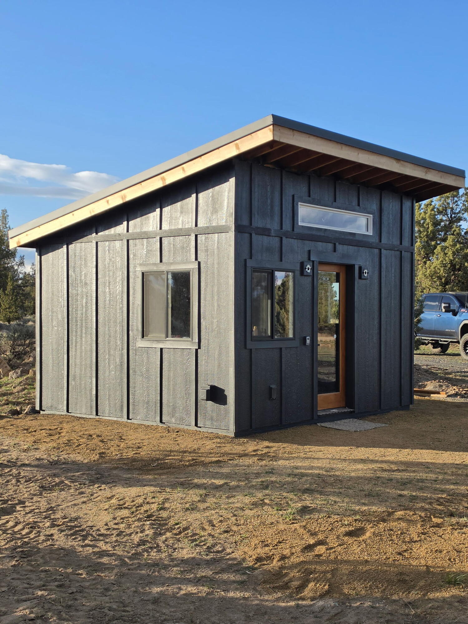 Compact dark-gray building with a single-slope roof and wood-accented glass entry on a bare lot.