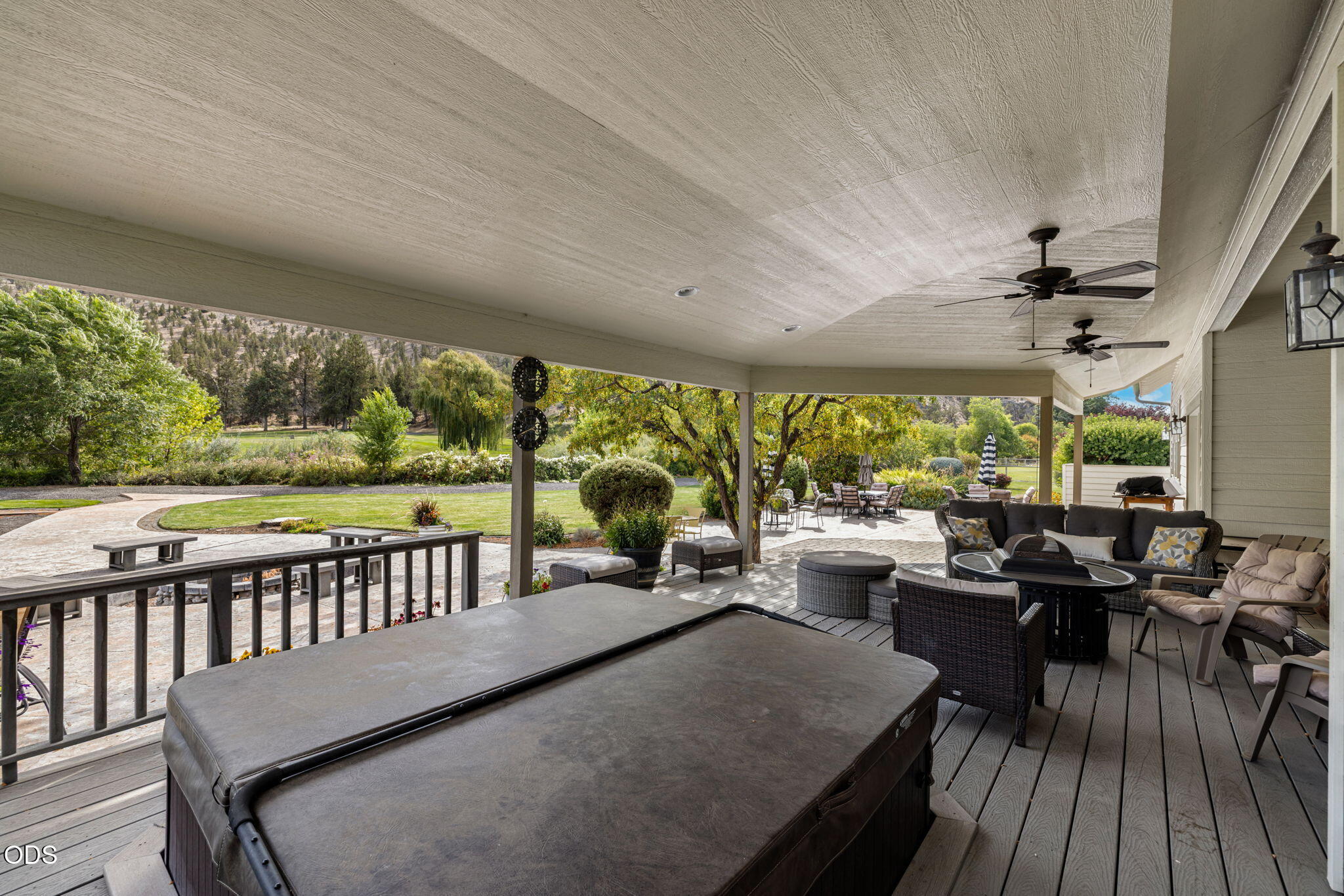 Covered deck with a large hot tub and lounge seating overlooking a landscaped yard and wooded hills.