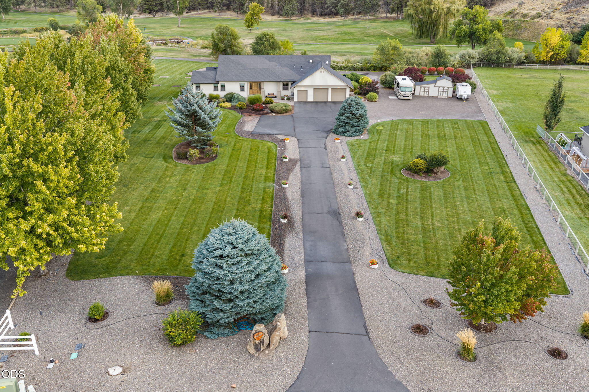 Aerial view of a landscaped home with a long driveway, striped lawn, RV, and small outbuildings.