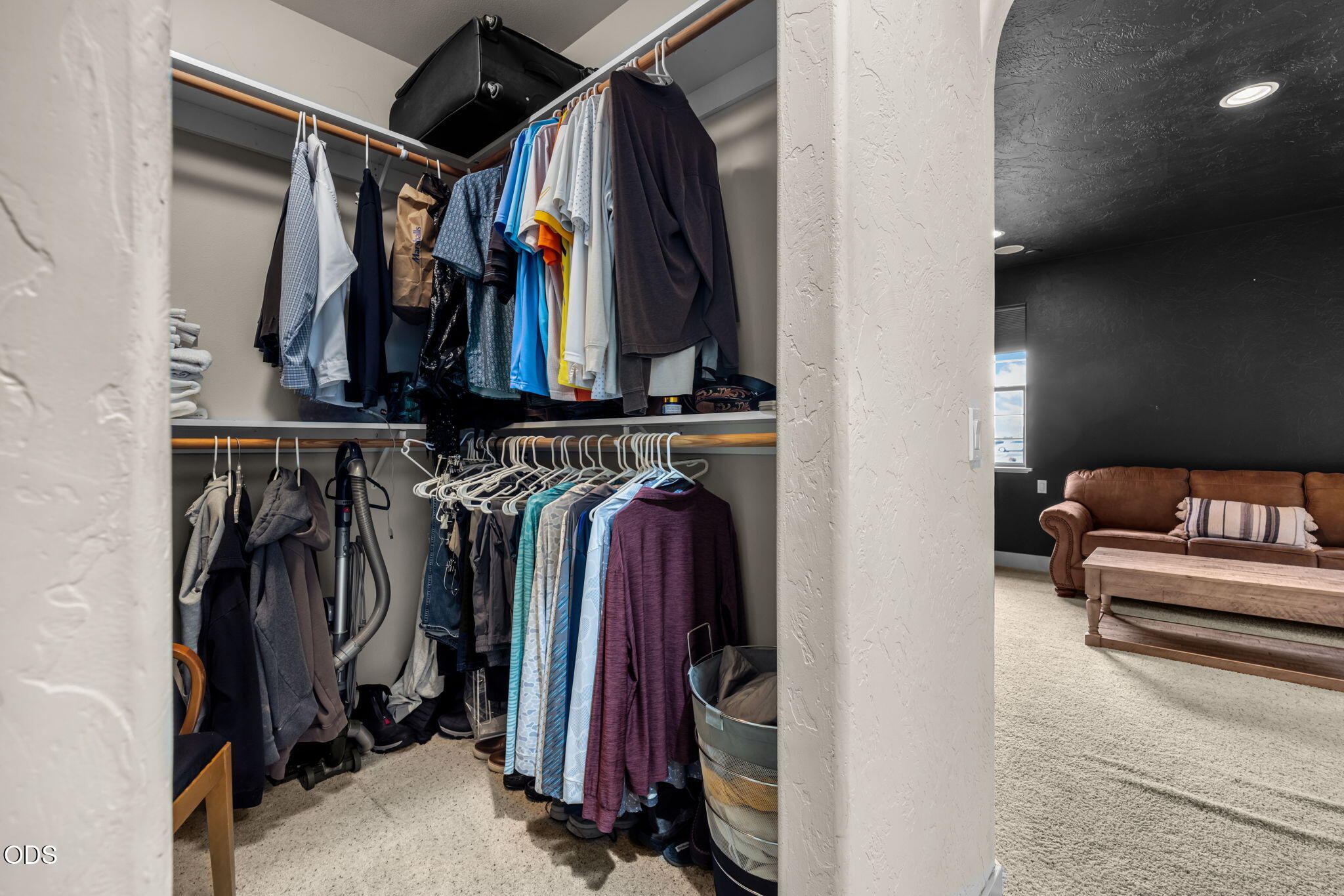 Walk-in closet packed with hanging clothes, opening onto a living room with a brown sofa.