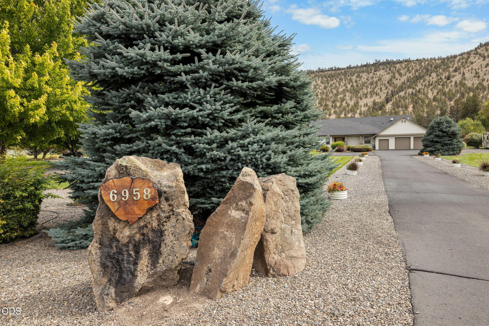 Single-story home with a three-car garage behind a large evergreen and rock address marker.