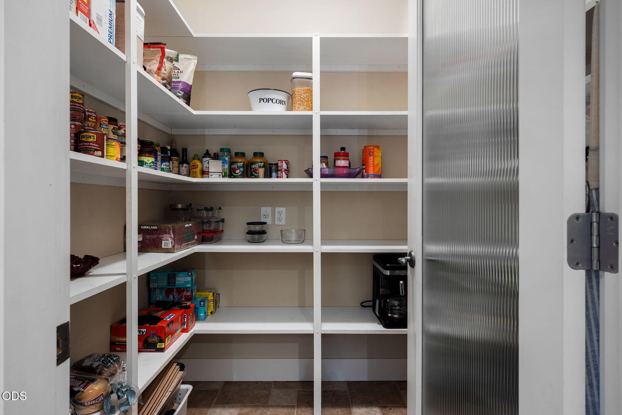 Walk-in pantry with wraparound white shelves stocked with food and a coffee maker.