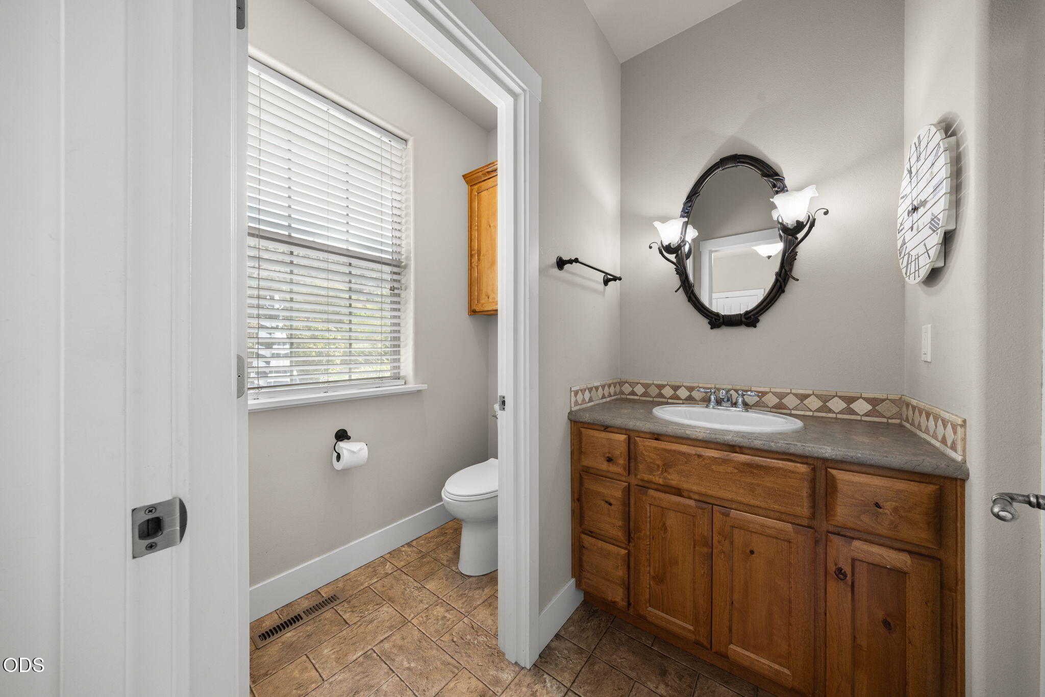 Bathroom with a wooden vanity and oval mirror, adjoining a separate toilet area with a window.