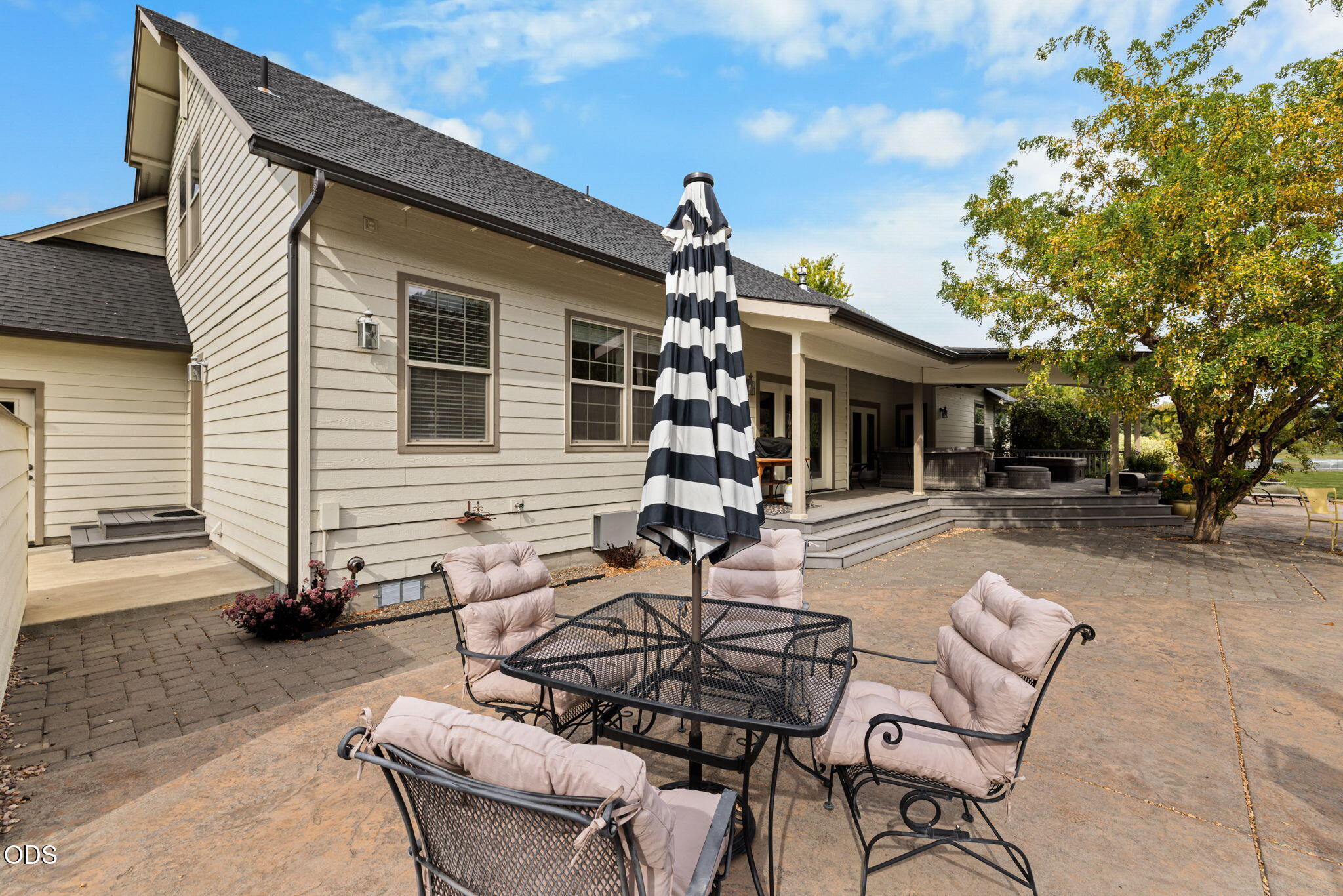Home’s rear patio with cushioned dining furniture and striped umbrella beside a covered deck.