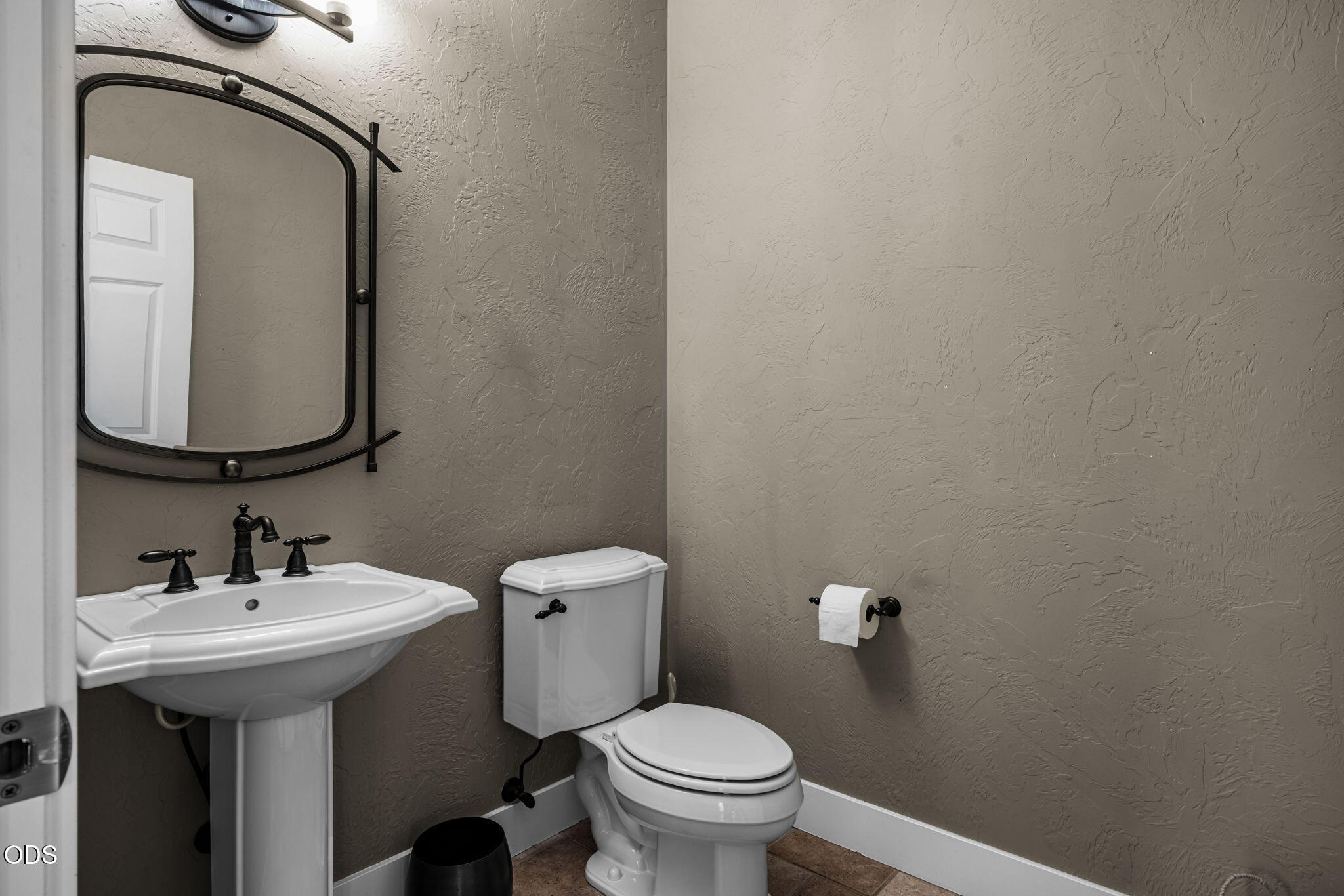 Bathroom with a pedestal sink, toilet, and large black-framed mirror against textured taupe walls.