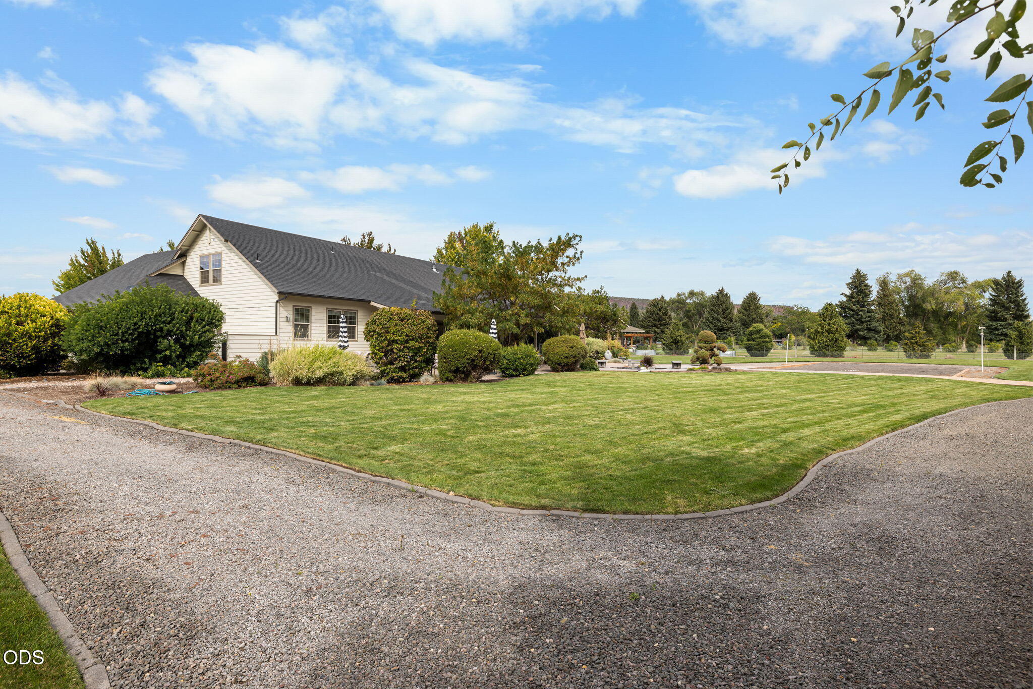 House with landscaped grounds, broad lawn, gravel driveway, and a paved recreation court.