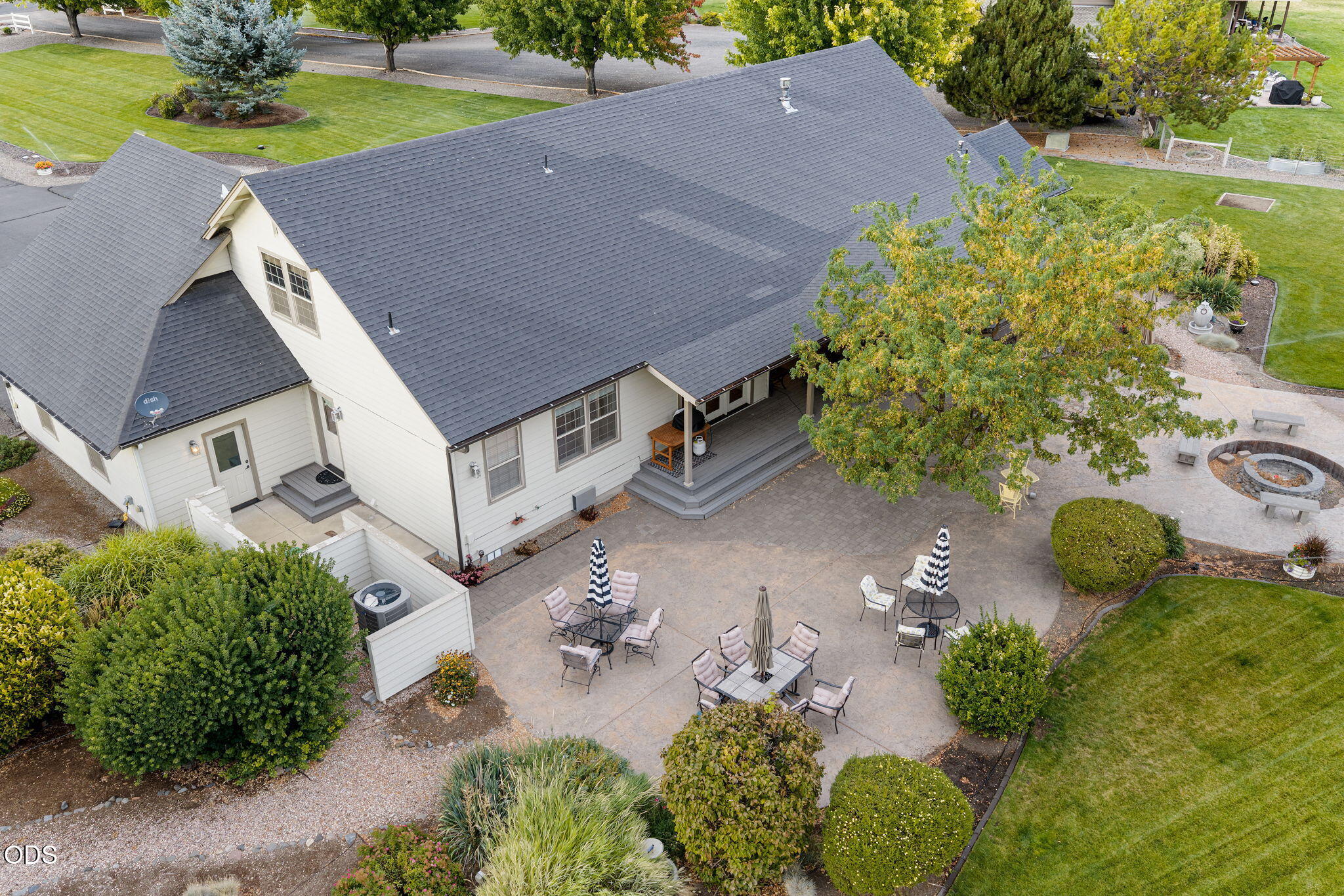 Aerial view of a home with a broad furnished patio, covered porch, circular fire pit, and landscaped yard.