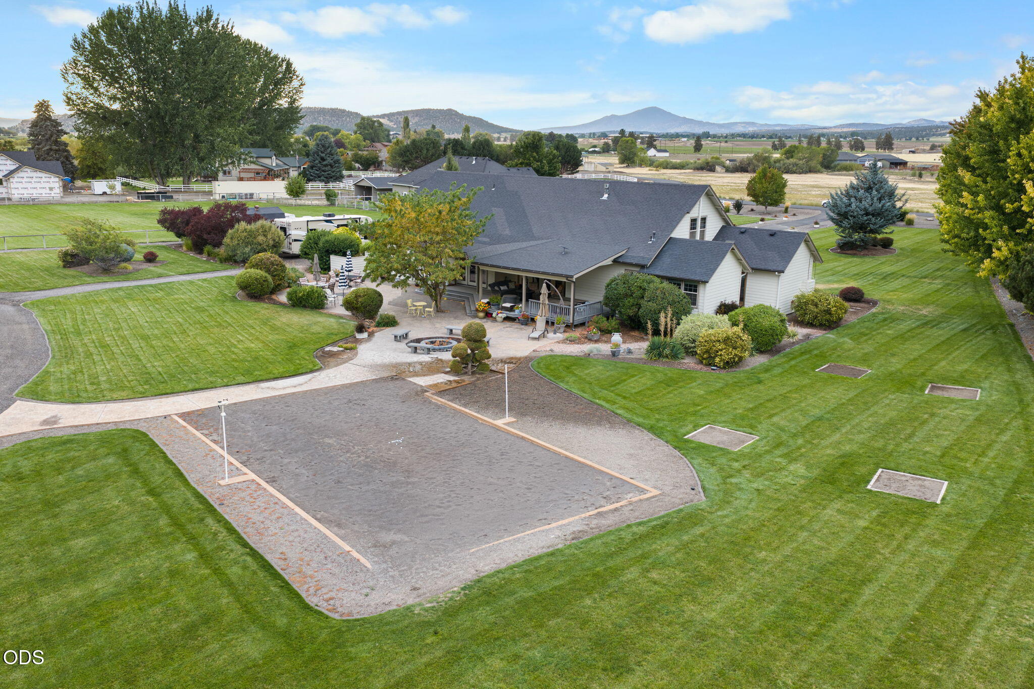 Aerial view of a home with a gravel volleyball court, landscaped lawn, and distant mountain views.