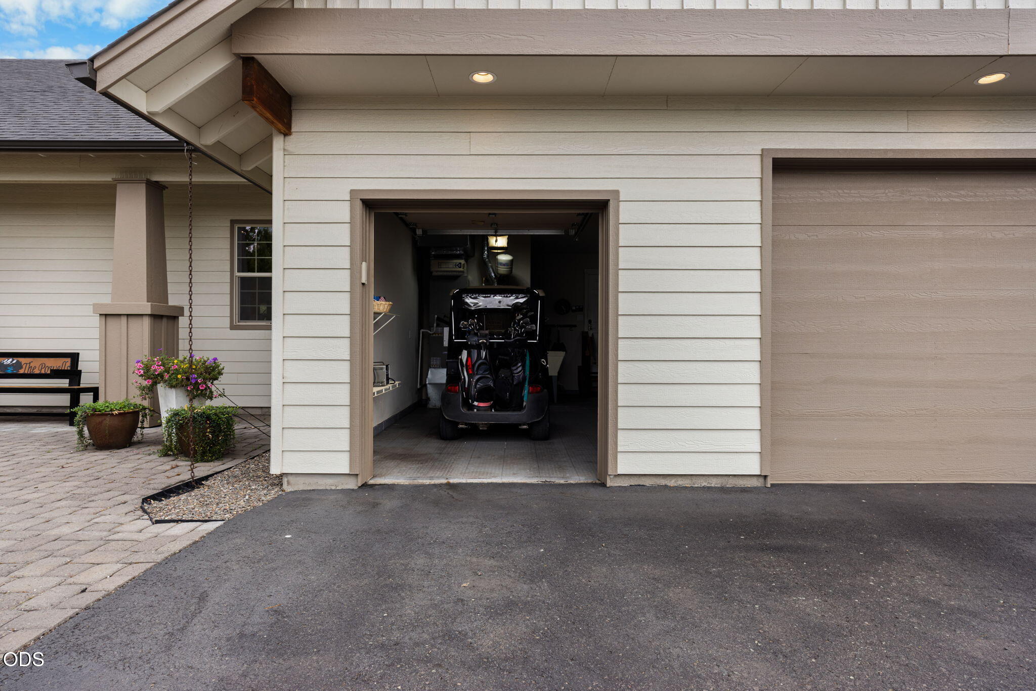 Open garage with a golf cart and golf bags, beside a paved driveway and flower planters.