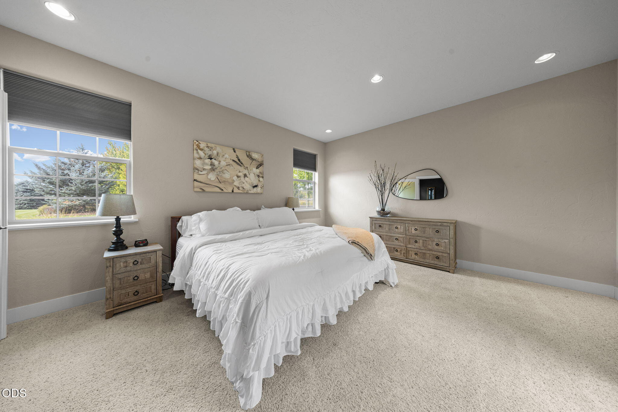 Bright bedroom with a white ruffled bed, wooden furniture, beige carpet, and two large windows.