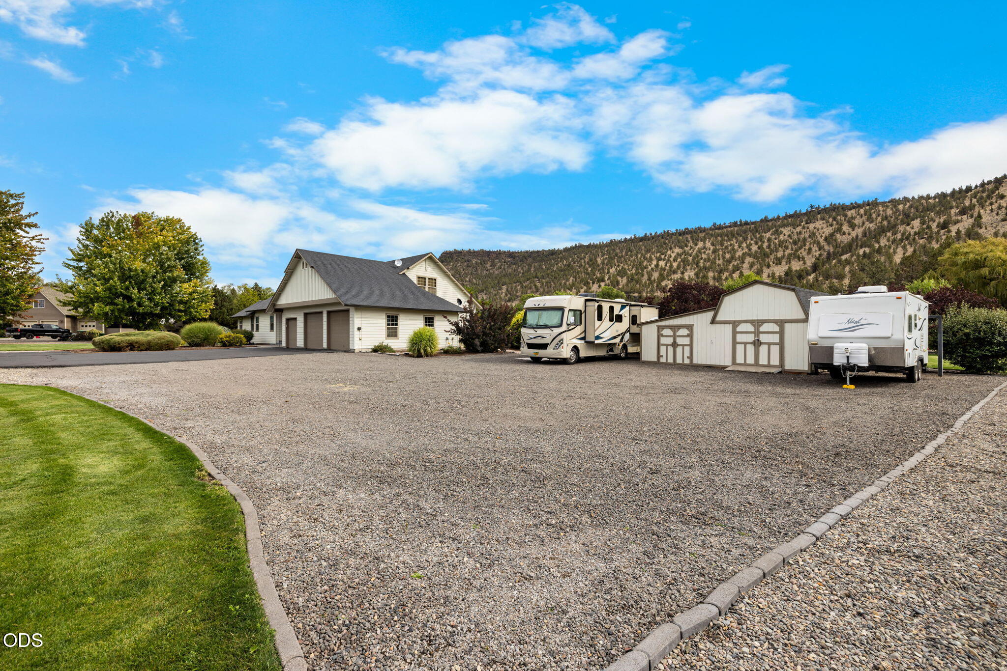 Home and detached garages beside a large gravel parking area with two RVs and a wooded hillside beyond.