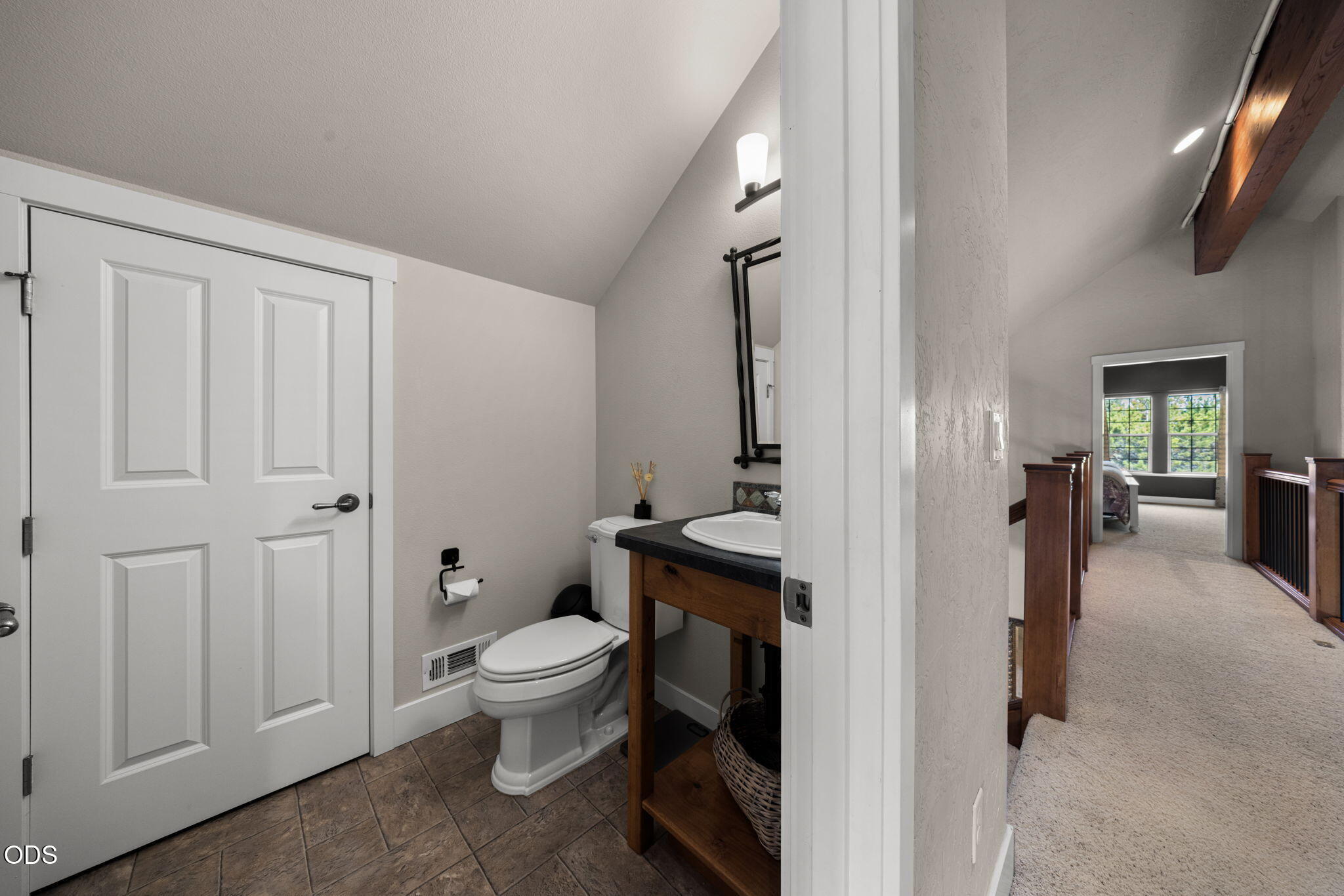 Bathroom with a wood vanity and tiled floor, opening onto a carpeted hallway with wood railings.