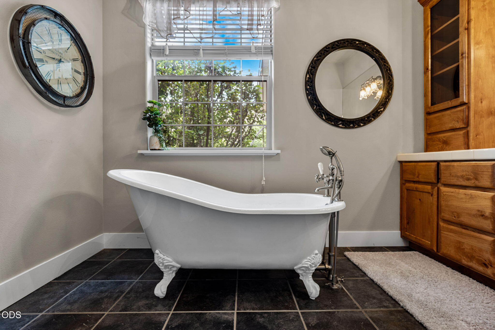 Bathroom with a white clawfoot tub beneath a window, beside a wood vanity and ornate round mirror.