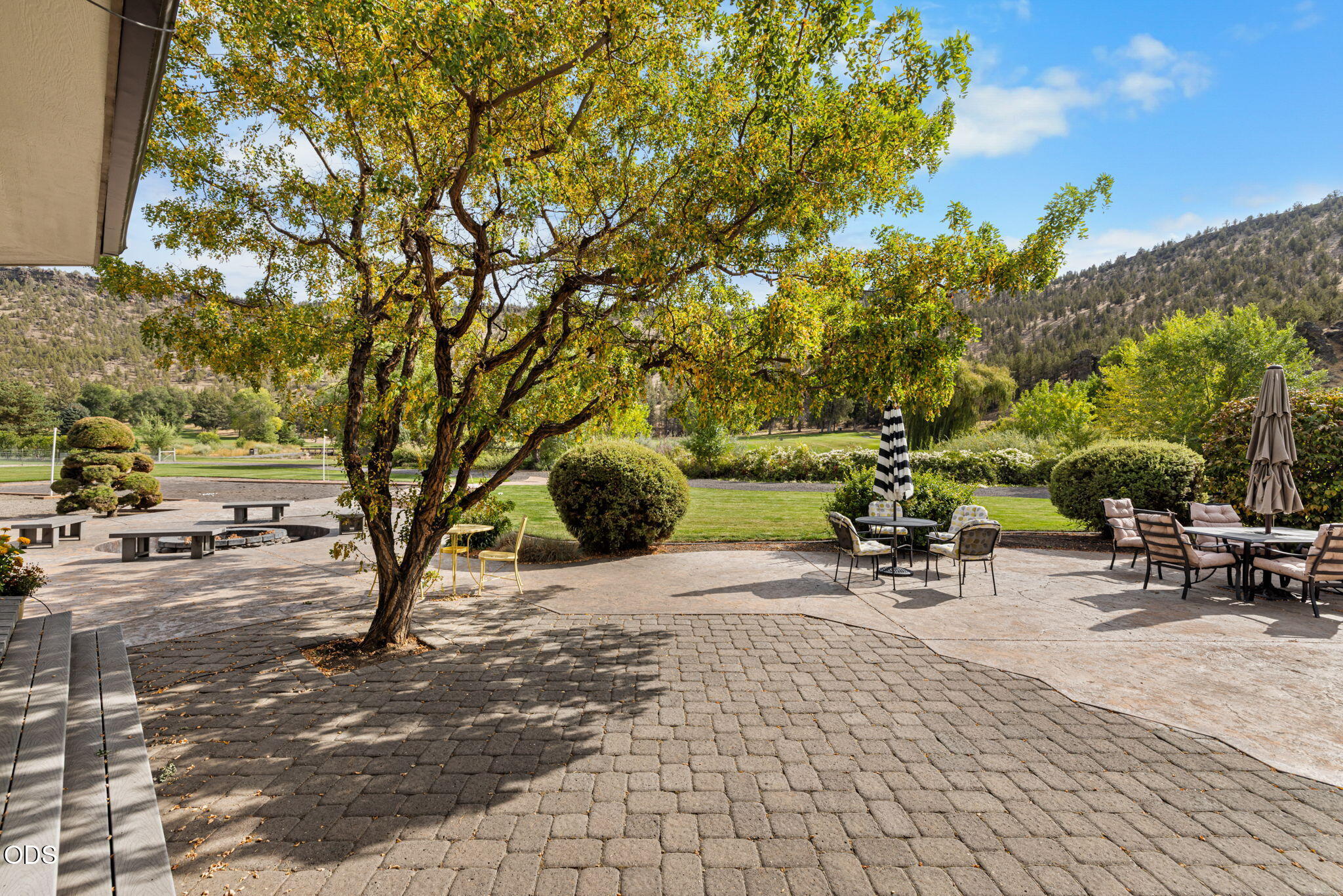 Paver patio with outdoor dining sets beneath a large tree, overlooking a lawn and wooded hills.