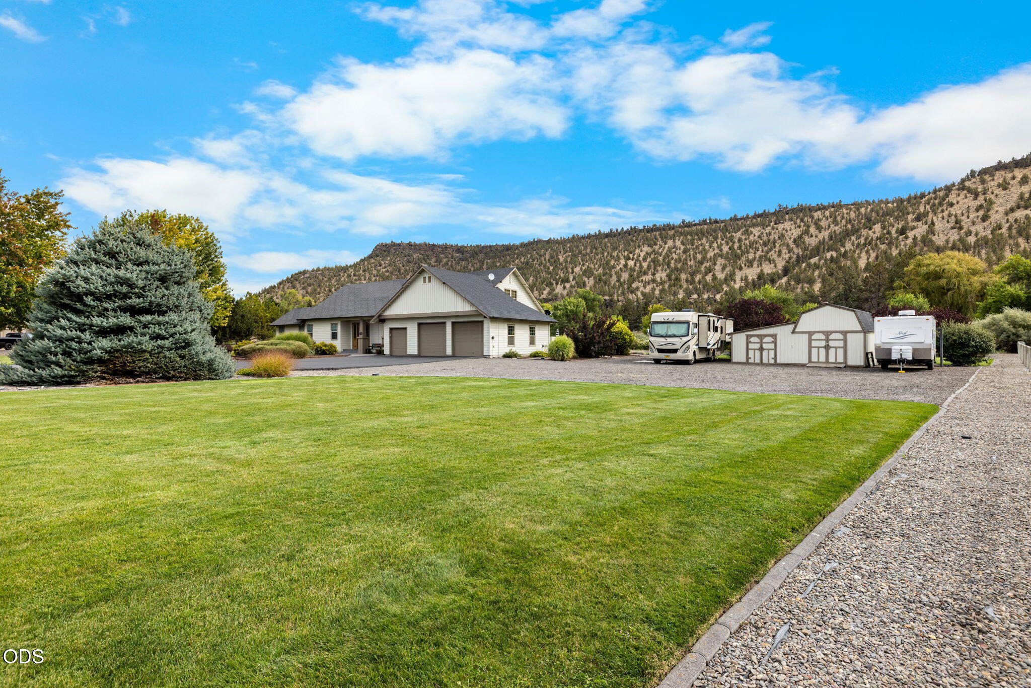 Home with a broad green lawn, attached garages, RVs, and a tree-covered hillside behind it.