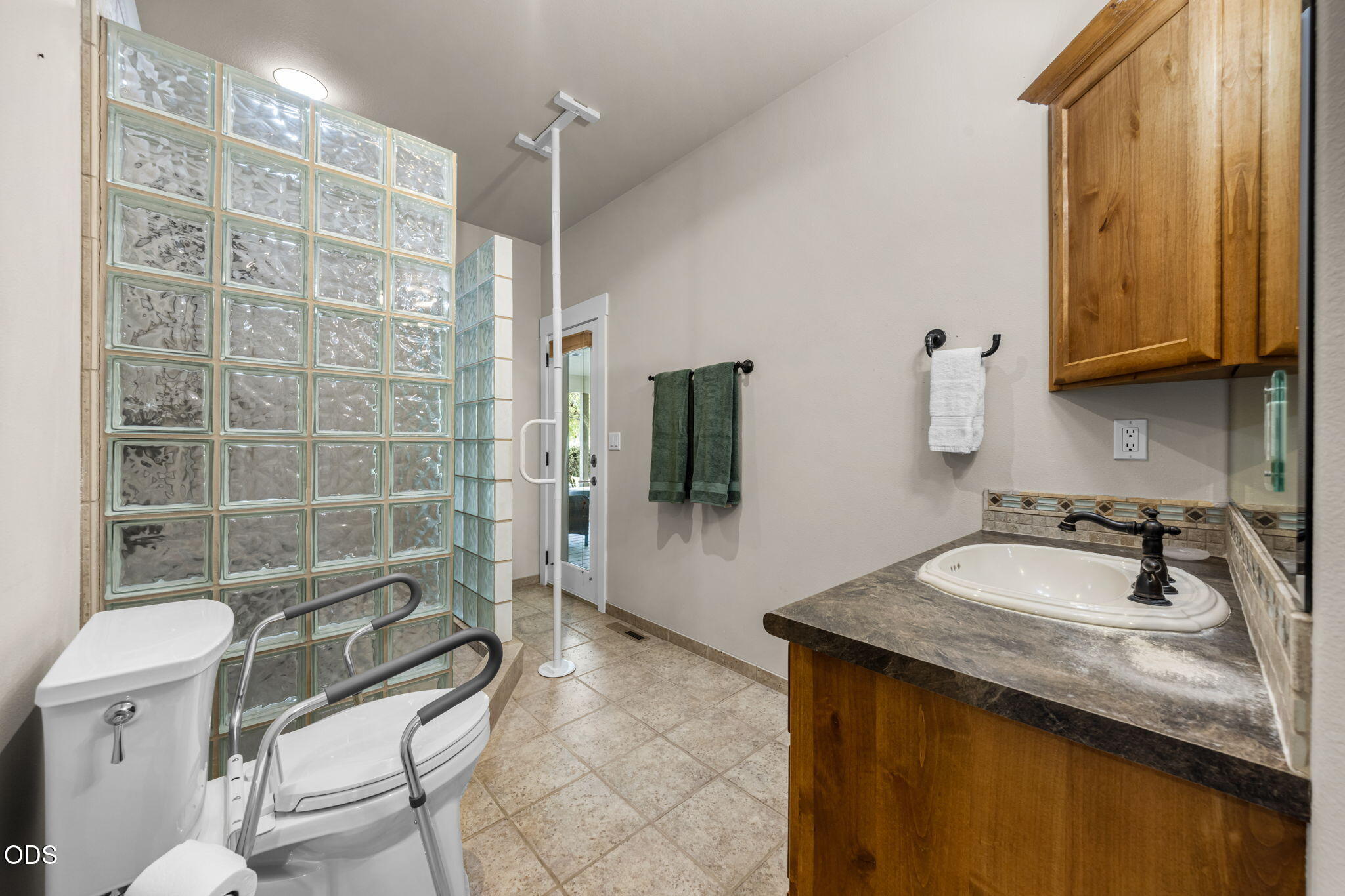 Bathroom with glass-block shower wall, toilet grab bars, and wood vanity with oval sink.