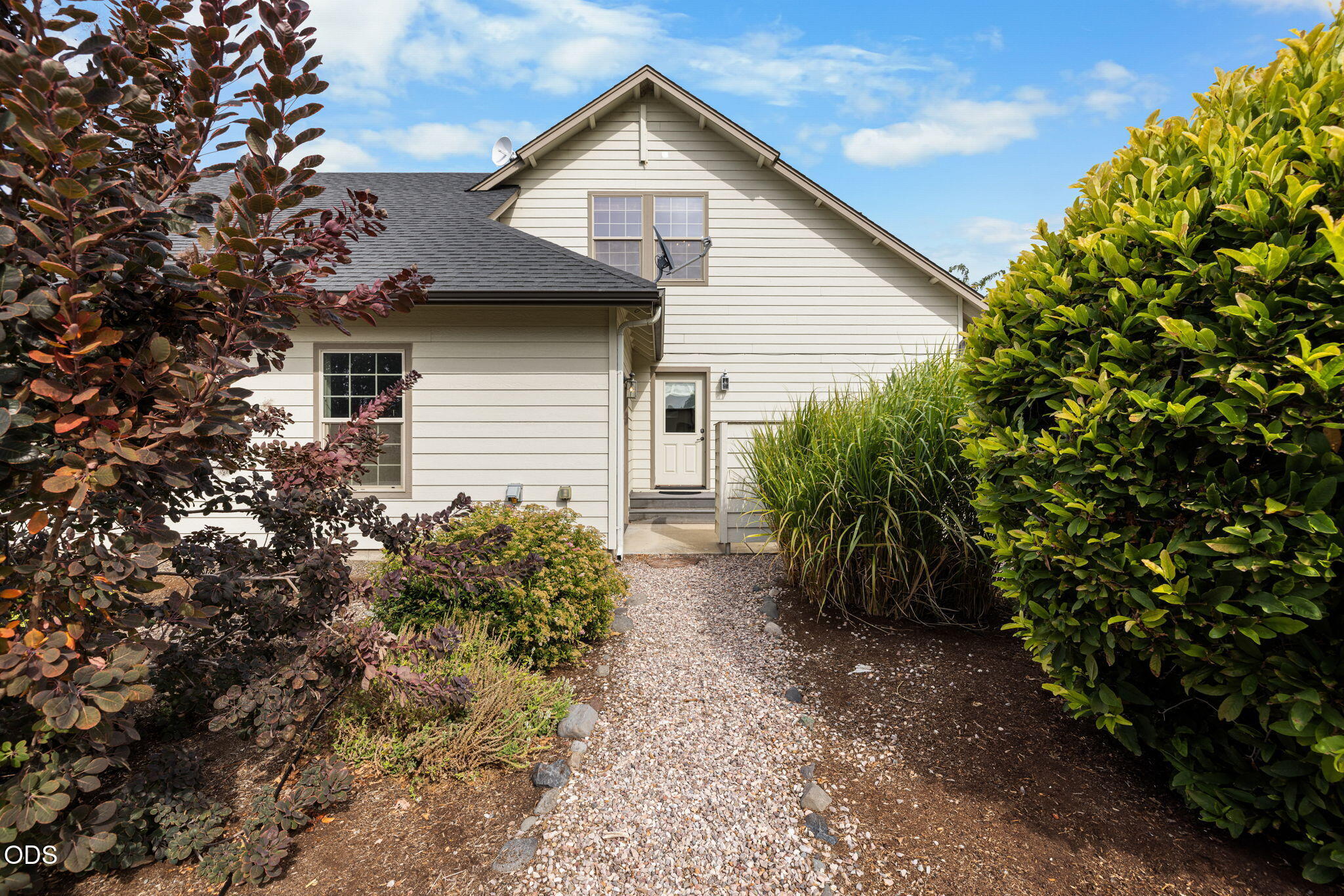 Light-colored two-story house with a gravel path to its side entrance, framed by dense landscaping.