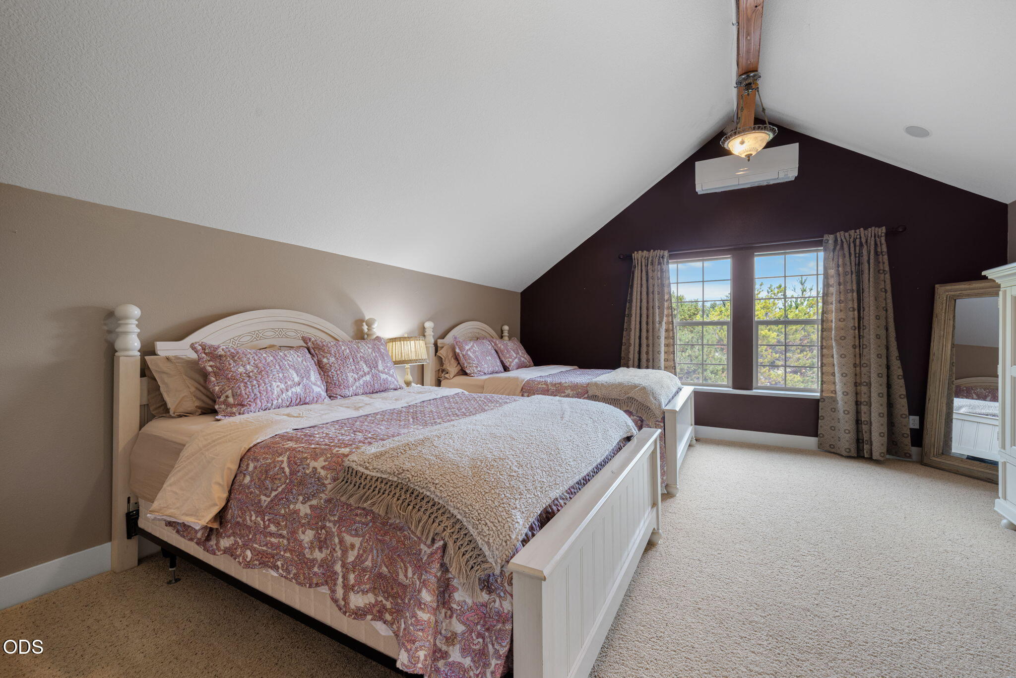 Bedroom with two patterned beds beneath a vaulted ceiling, beside curtained windows overlooking trees.