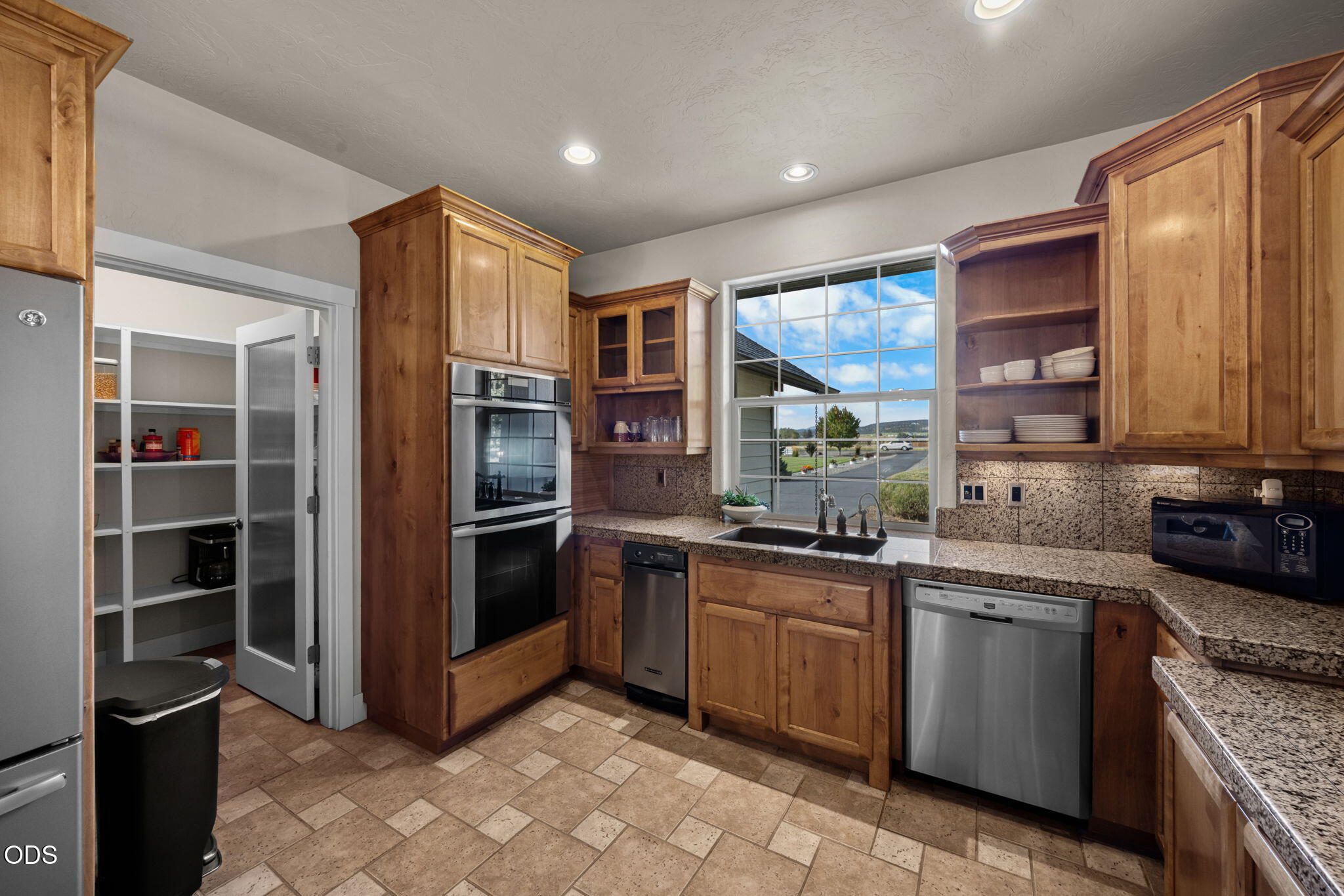 Kitchen with warm wood cabinets, granite counters, double ovens, and a large window above the sink.