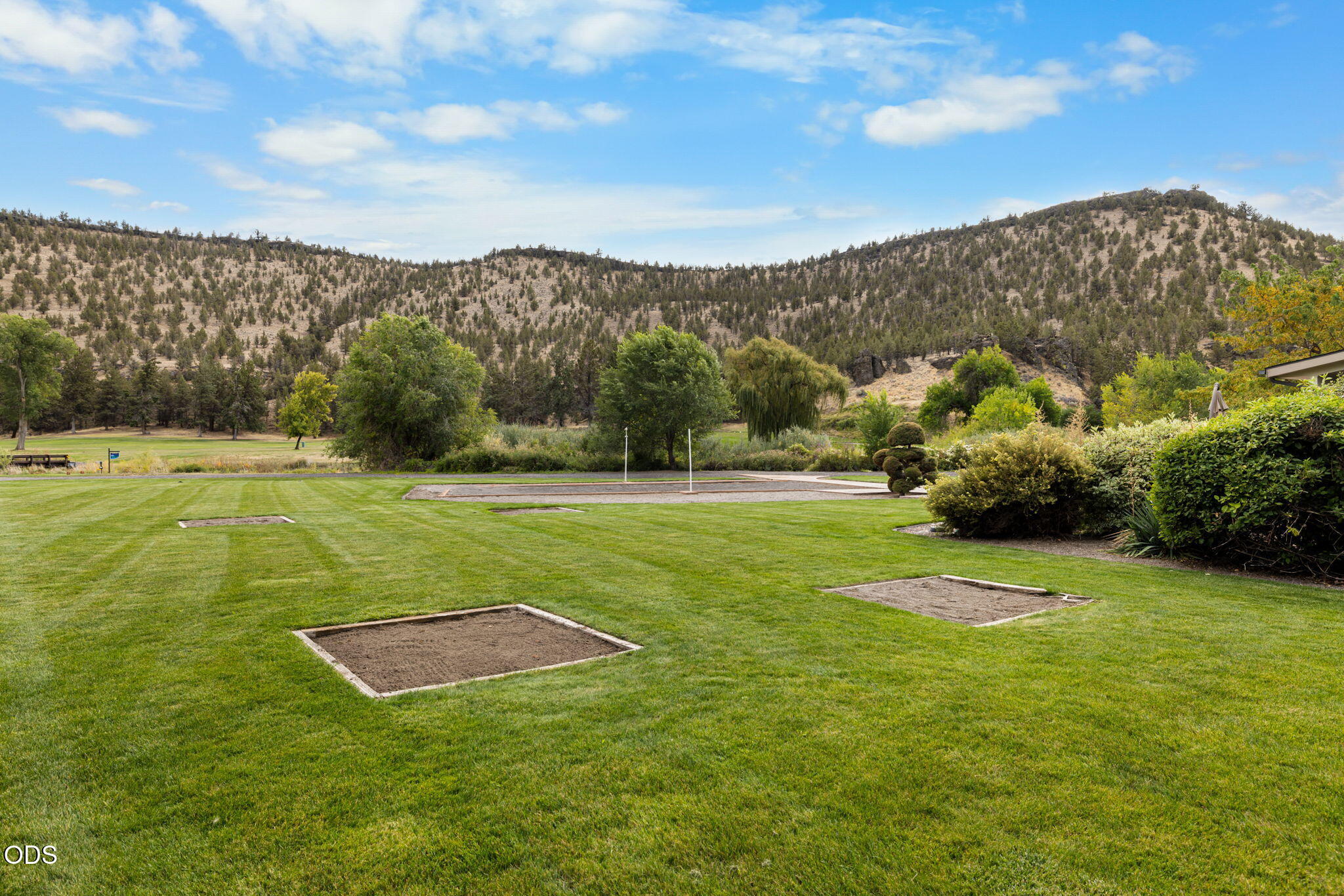 Lawn with sand pits and a gravel recreation area below tree-covered hills.