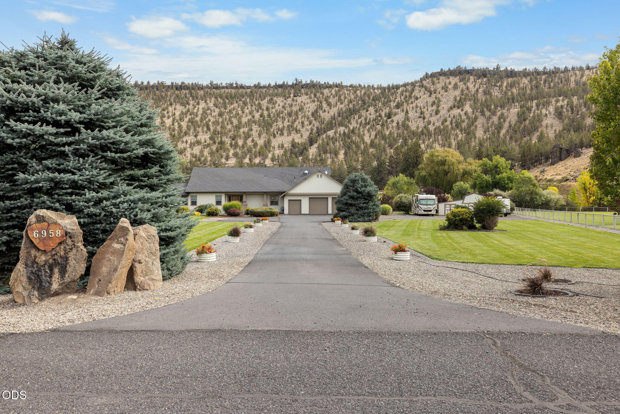 Single-story home with a two-car garage, long landscaped driveway, and tree-dotted hillside behind it.