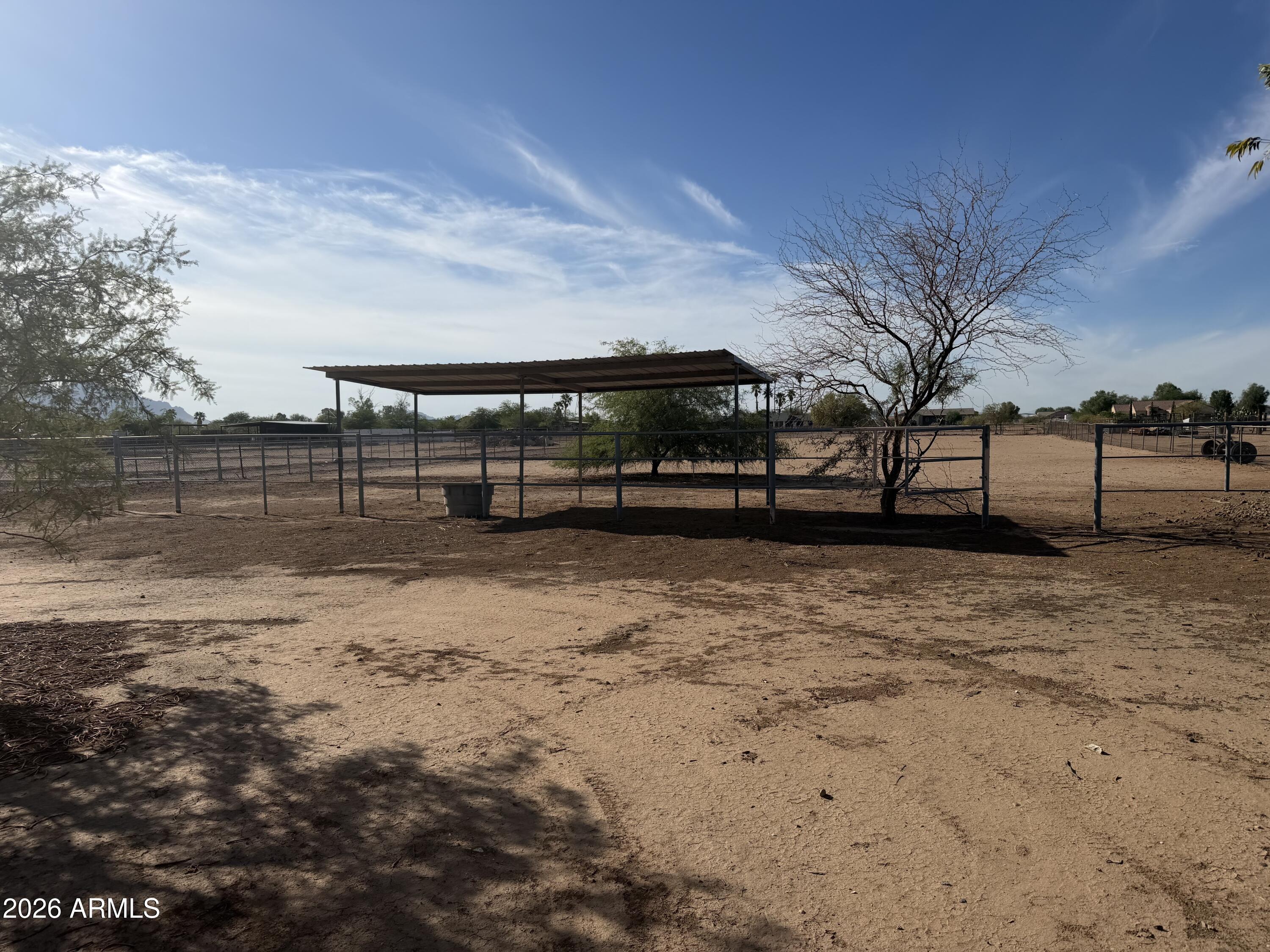 Fenced dirt paddock with a metal shade shelter, water trough, and sparse trees.