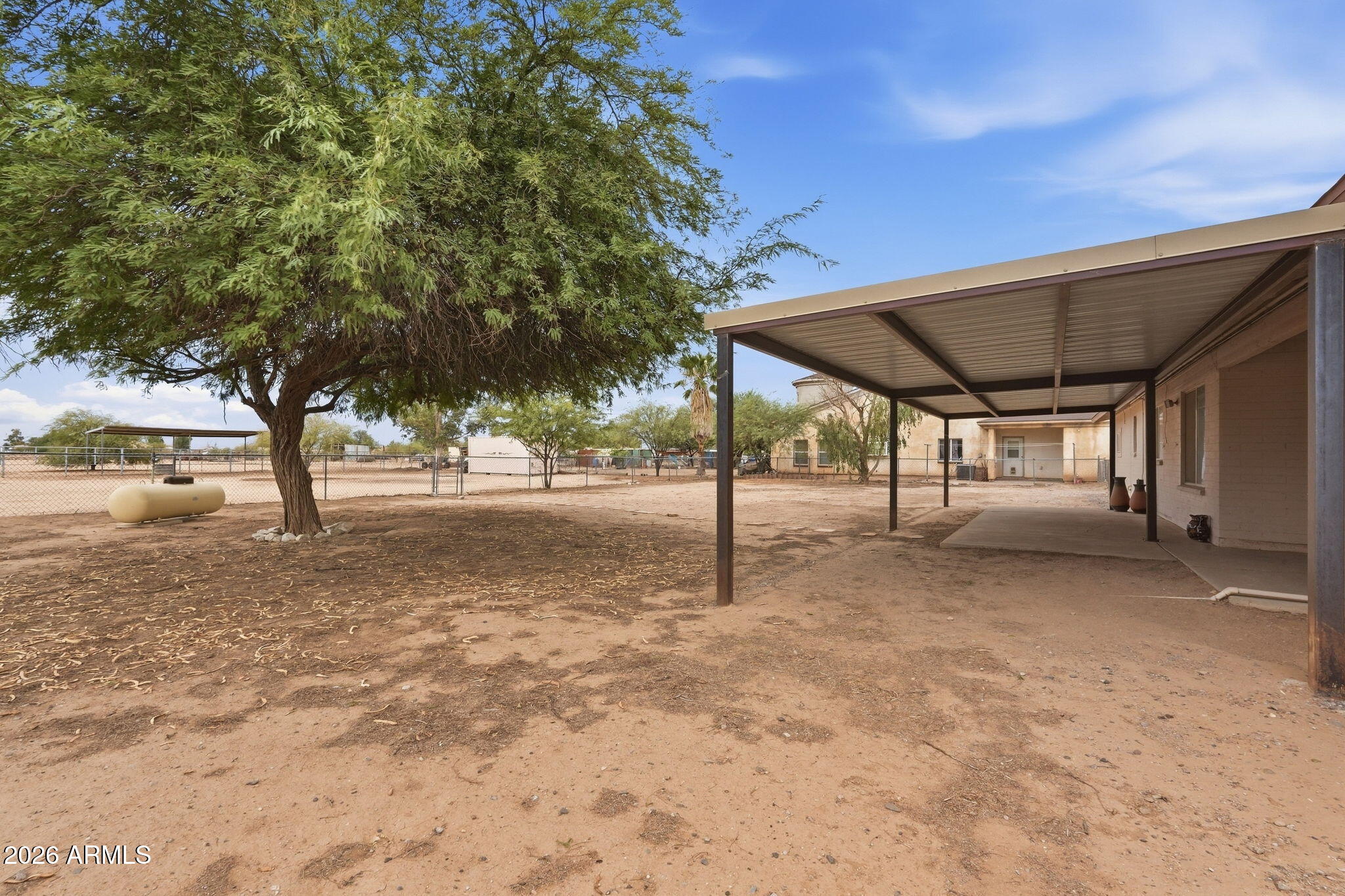 Home exterior with a broad covered patio, large shade tree, and fenced dirt yard.