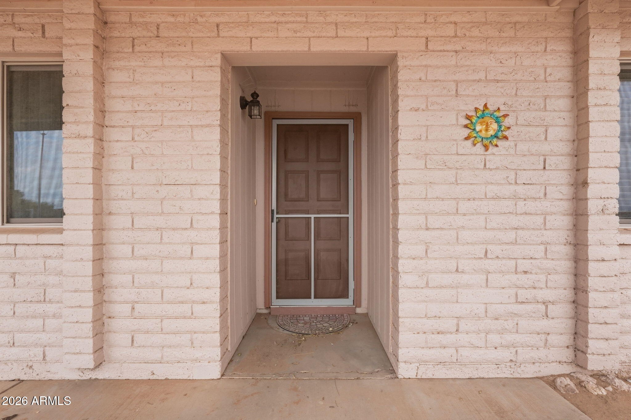 Recessed brown front door with a screen door in a pale block exterior, beside a colorful sun decoration.