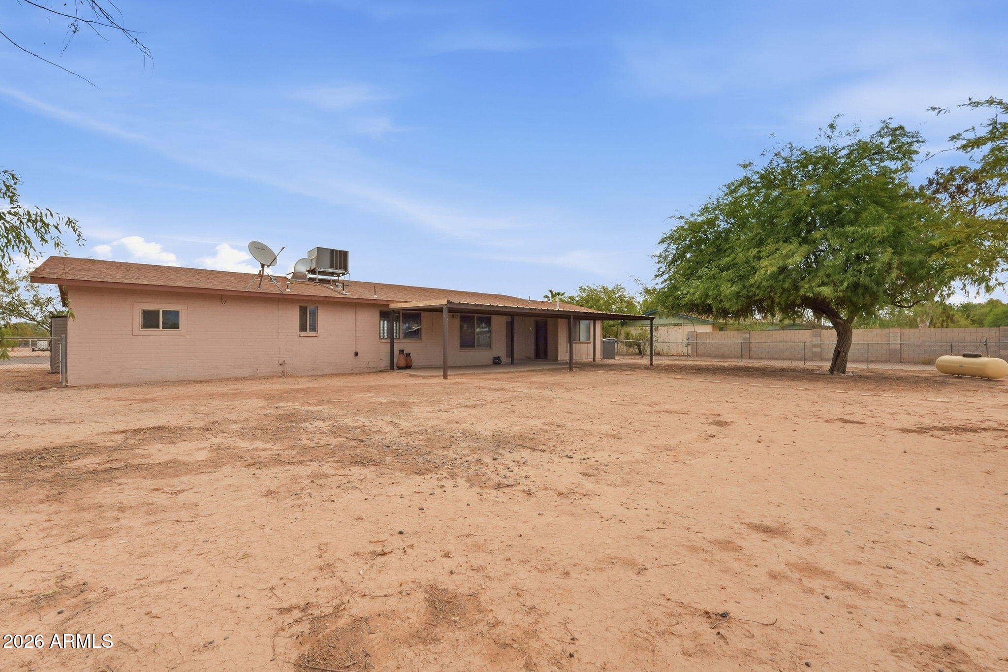 Single-story home's rear exterior with covered patio, fenced dirt yard, and mature tree.