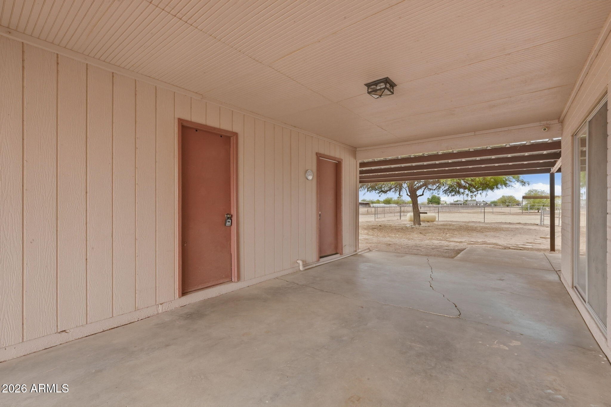 Covered concrete patio with two doors opening toward a fenced yard and shade tree.