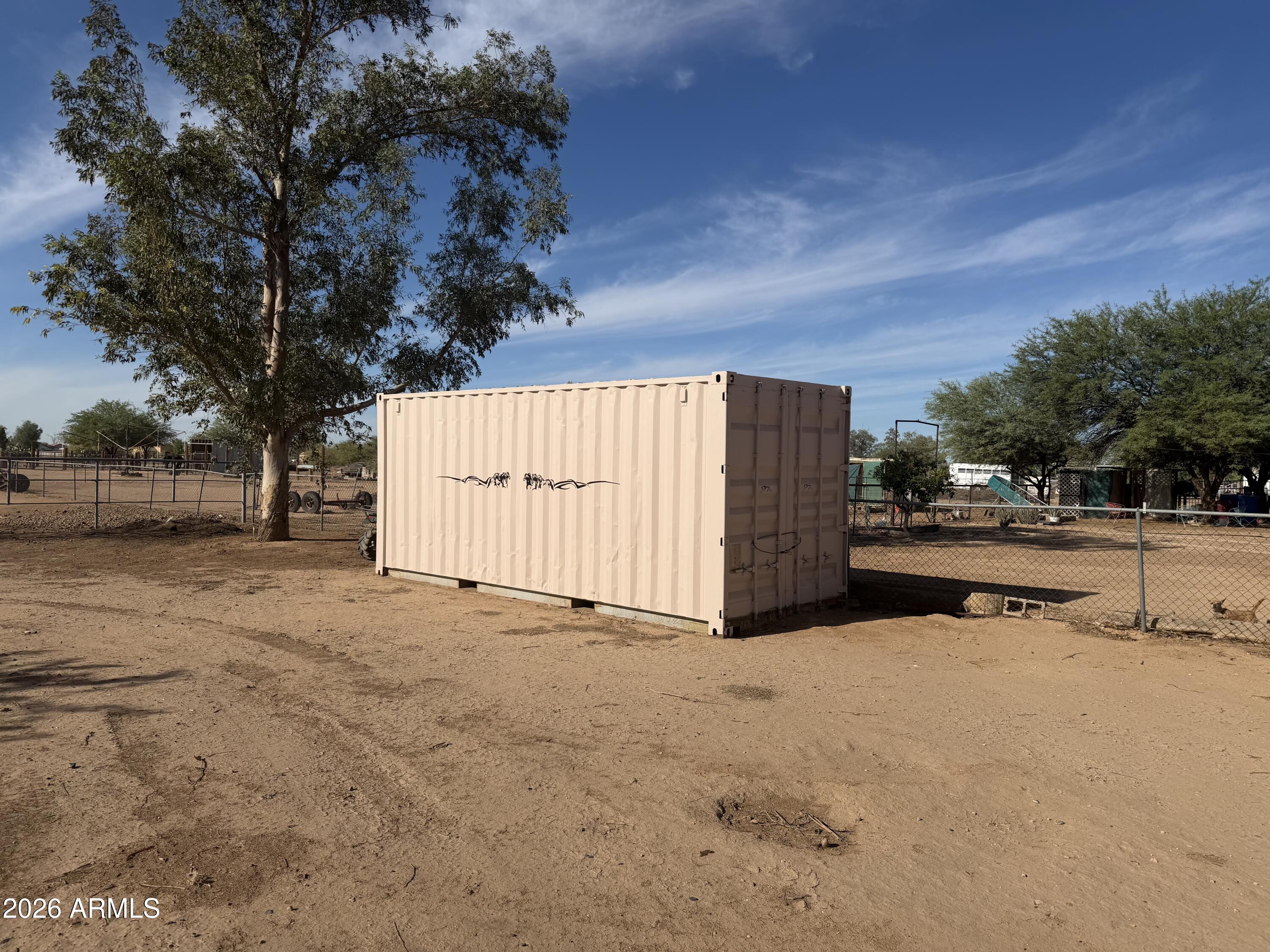 Shipping container in a fenced dirt yard beneath mature trees.