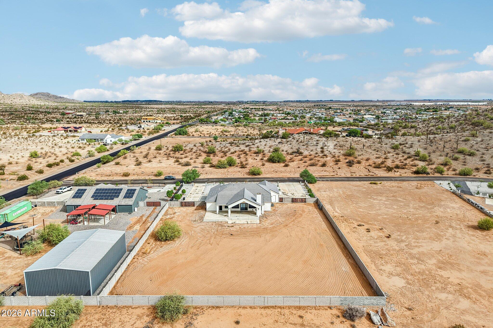 Aerial view of a desert property with a walled lot, solar-panelled outbuilding, and distant rocky hills.